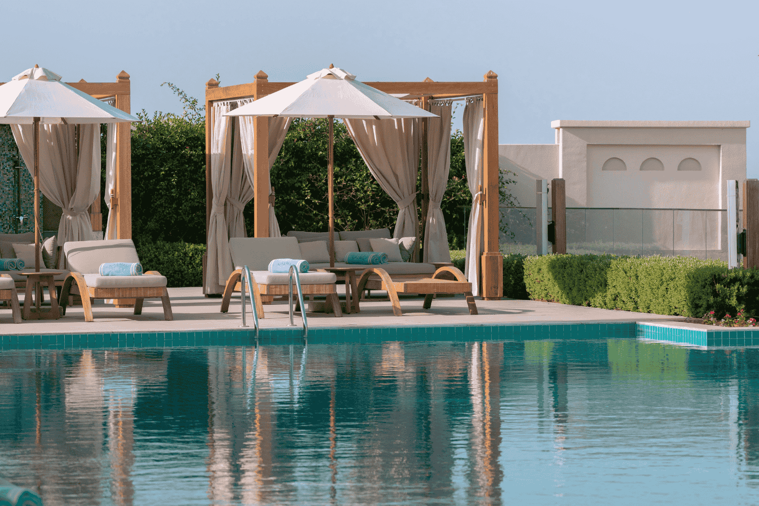 A serene swimming pool area with lounge chairs, beige umbrellas, shaded cabanas, and neatly trimmed bushes against a light blue sky.