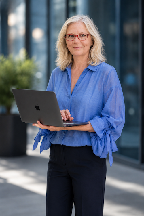 A woman with shoulder-length gray hair and glasses, wearing a blue blouse, stands outdoors holding a silver laptop.