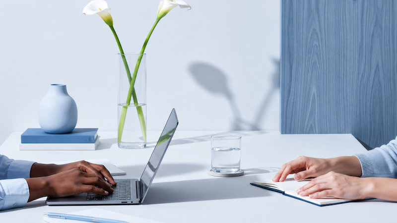 Two people sitting across from each other at a white table, one using a laptop and the other with a notepad. On the table are a glass of water, a vase with white calla lilies, and a small blue vase on top of two books. The background features a light wall and a blue wooden panel.