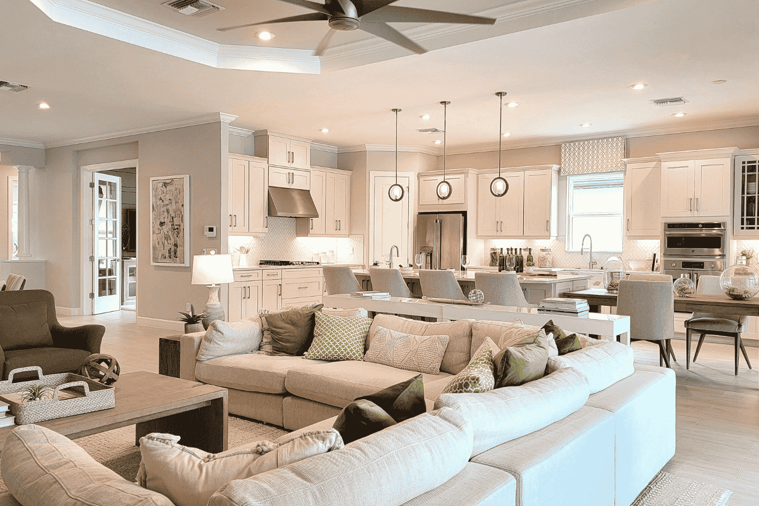 Open-concept living room and kitchen with beige sofa, pillows, armchair, kitchen island with chairs, white cabinetry, stainless steel appliances, pendant lights, and large windows.