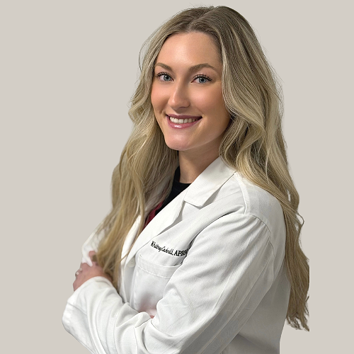Young woman with long, wavy blonde hair smiling, wearing a white lab coat with embroidered name and title, standing against a plain wall.