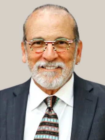 Smiling elderly man with glasses, a gray and white beard, and an earring, wearing a dark suit, white shirt, and a patterned tie.