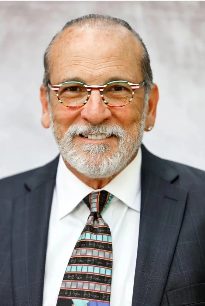 Smiling elderly man with glasses, a gray and white beard, and an earring, wearing a dark suit, white shirt, and a patterned tie.