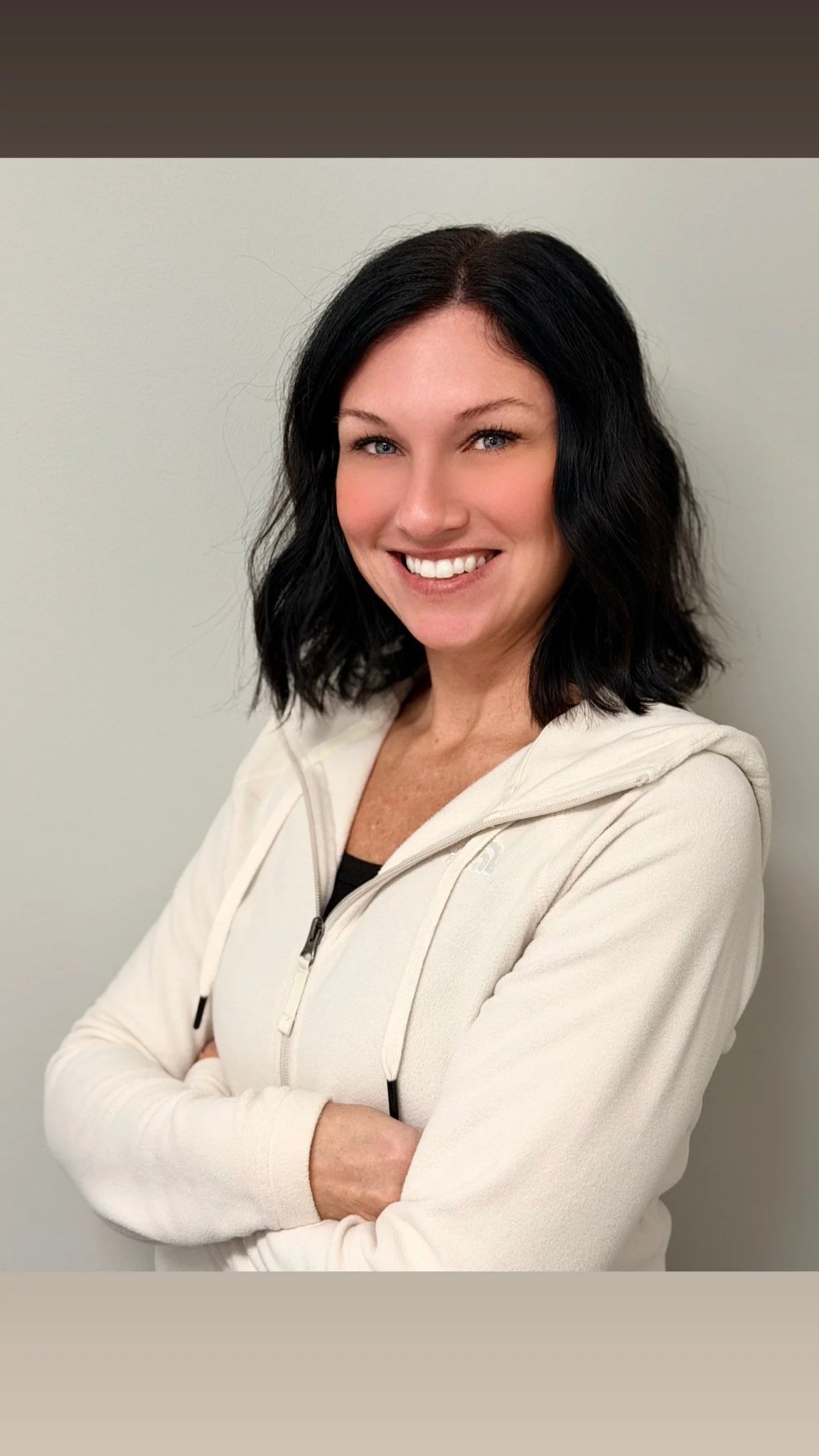 A smiling woman with shoulder-length black hair, crossed arms, wearing a white zip-up hoodie in front of a plain light-colored wall.