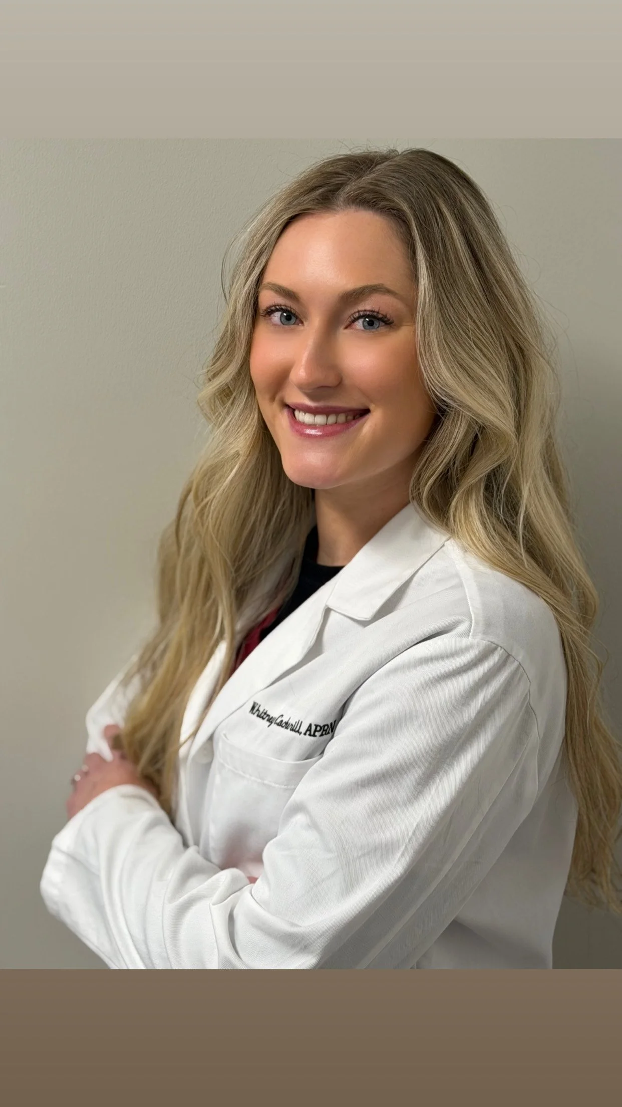 Young woman with long, wavy blonde hair smiling, wearing a white lab coat with embroidered name and title, standing against a plain wall.