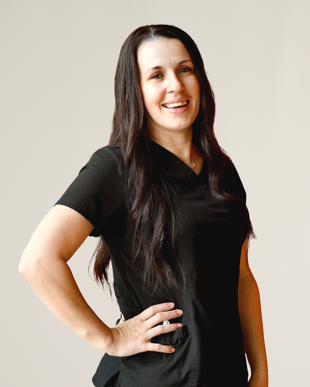 A woman with long black hair, smiling, wearing a black short-sleeve shirt, standing with her left hand on her hip against a plain light-colored background.