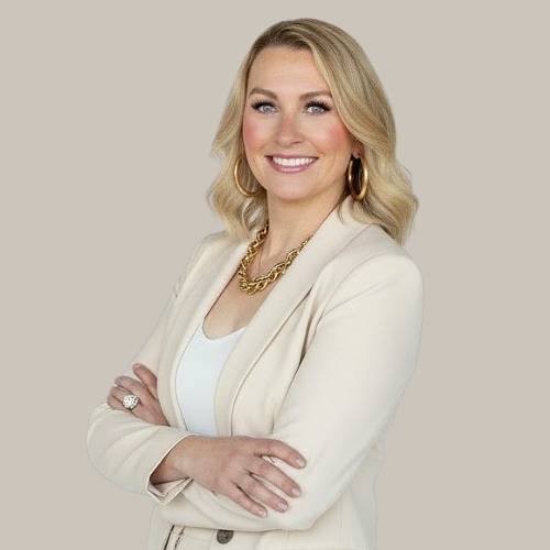 A smiling female healthcare professional wearing a white coat and teal scrubs, standing against a plain light-colored background.
