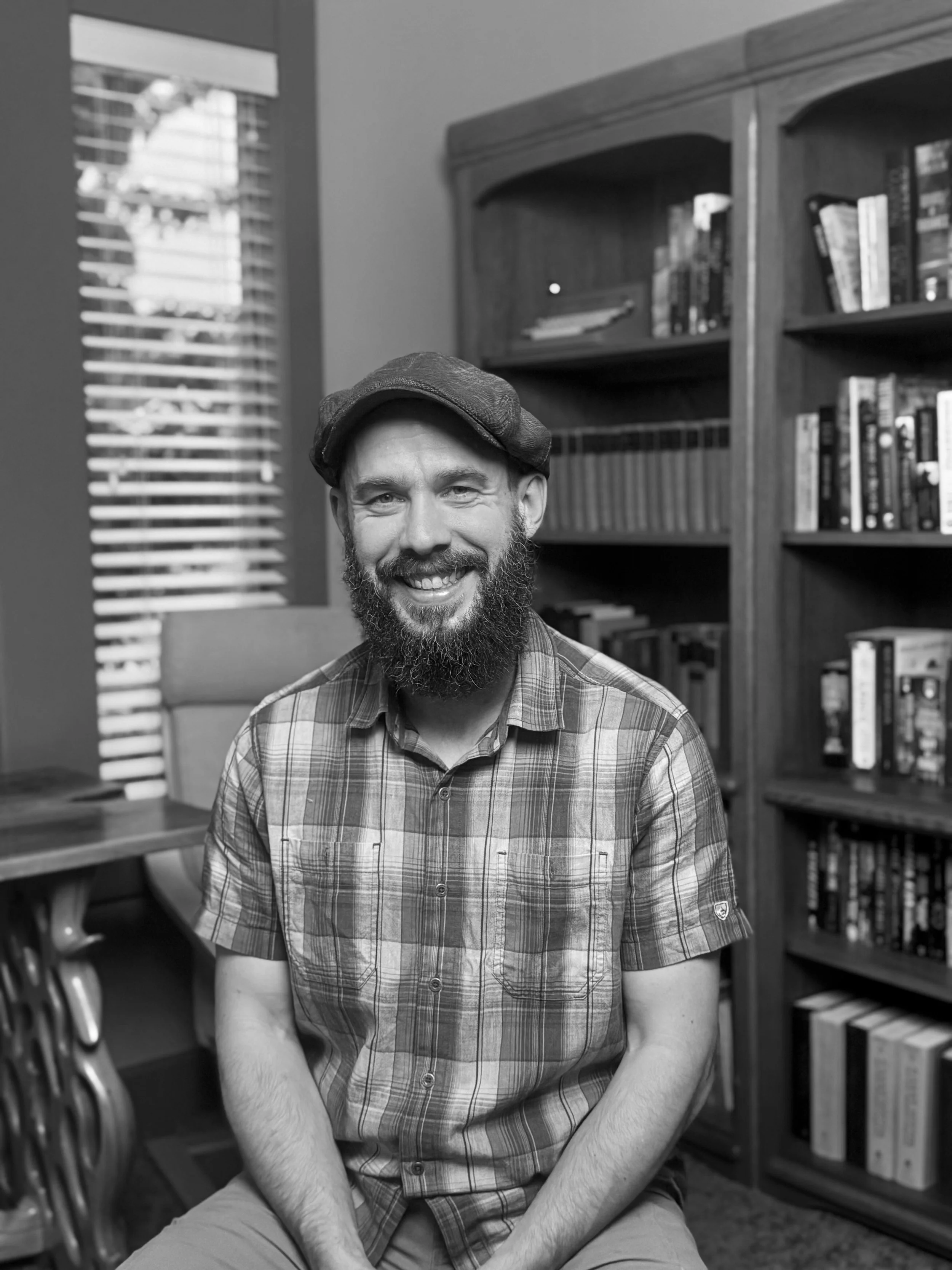 A black-and-white portrait of author Kaleb Erickson sitting in his office, with a softly blurred background of books on wooden shelves.
