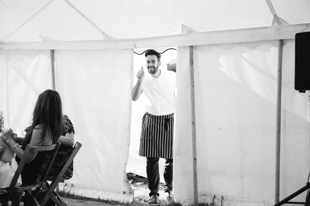A smiling male chef in a striped apron giving a thumbs-up as he stands in a marquee tent entrance, with a woman seated at a table visible nearby.
