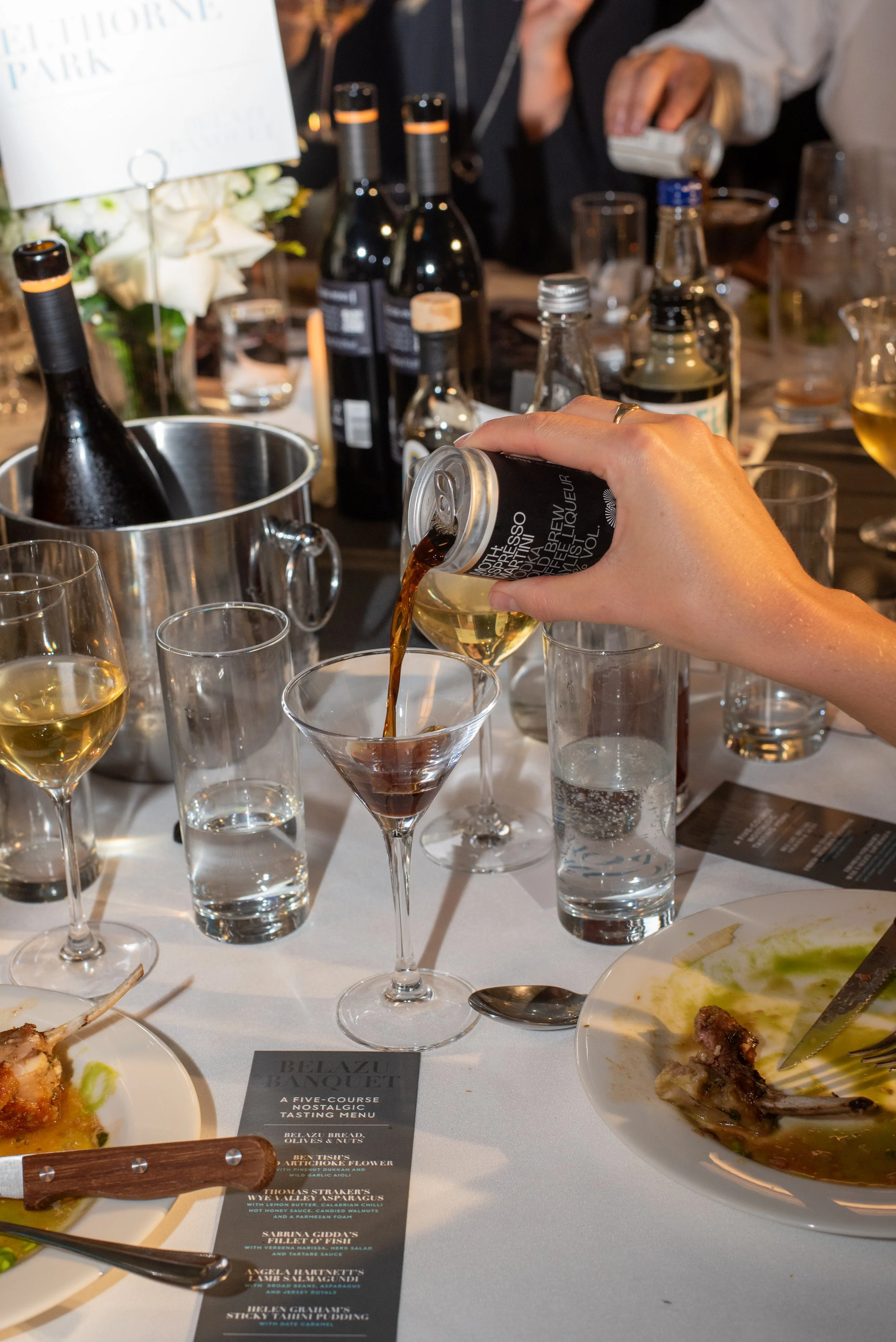 Person pouring a moth canned cocktail into a martini glass on a table set for a charity banquet, with empty and partially filled glasses, wine bottles, and plates of food around.