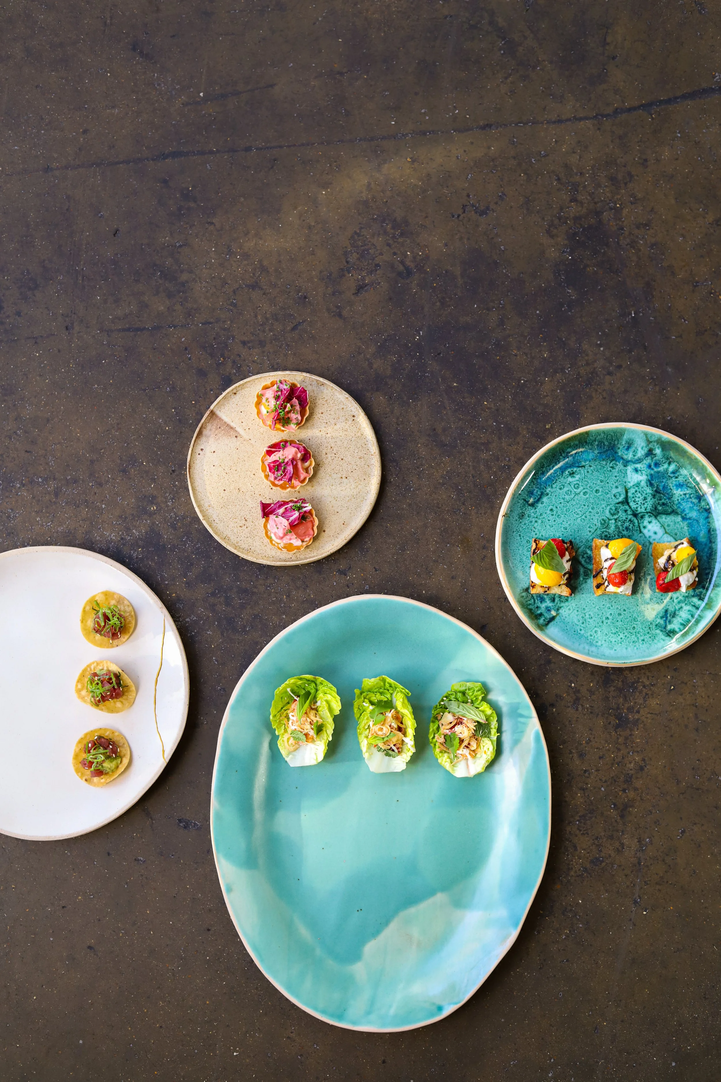 An overhead view of four colorful plates with small, artfully arranged canapes on an industrial surface.