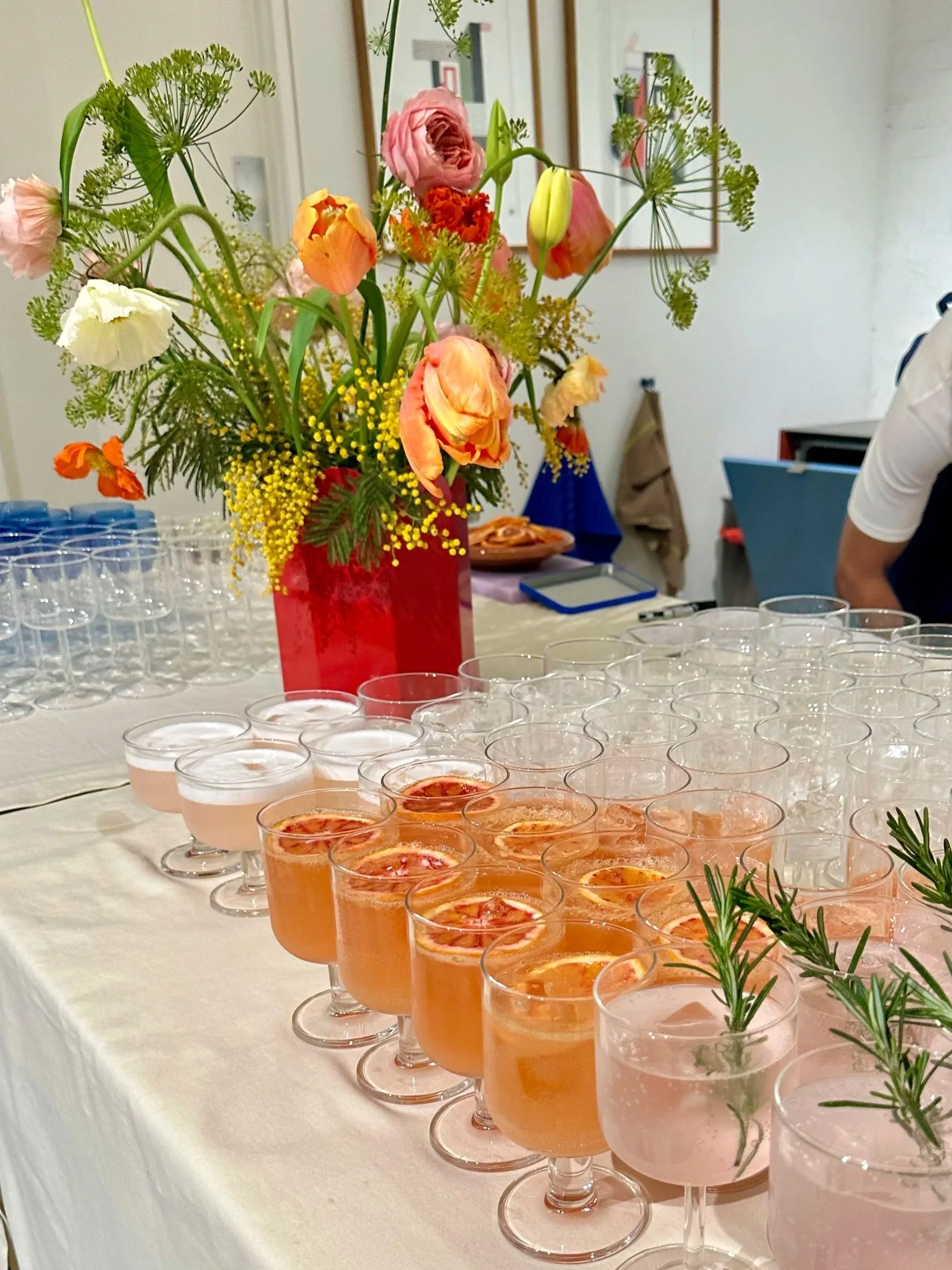 A table with a large floral arrangement in a red vase and multiple glasses of pink beverages garnished with lemon slices and herbs.
