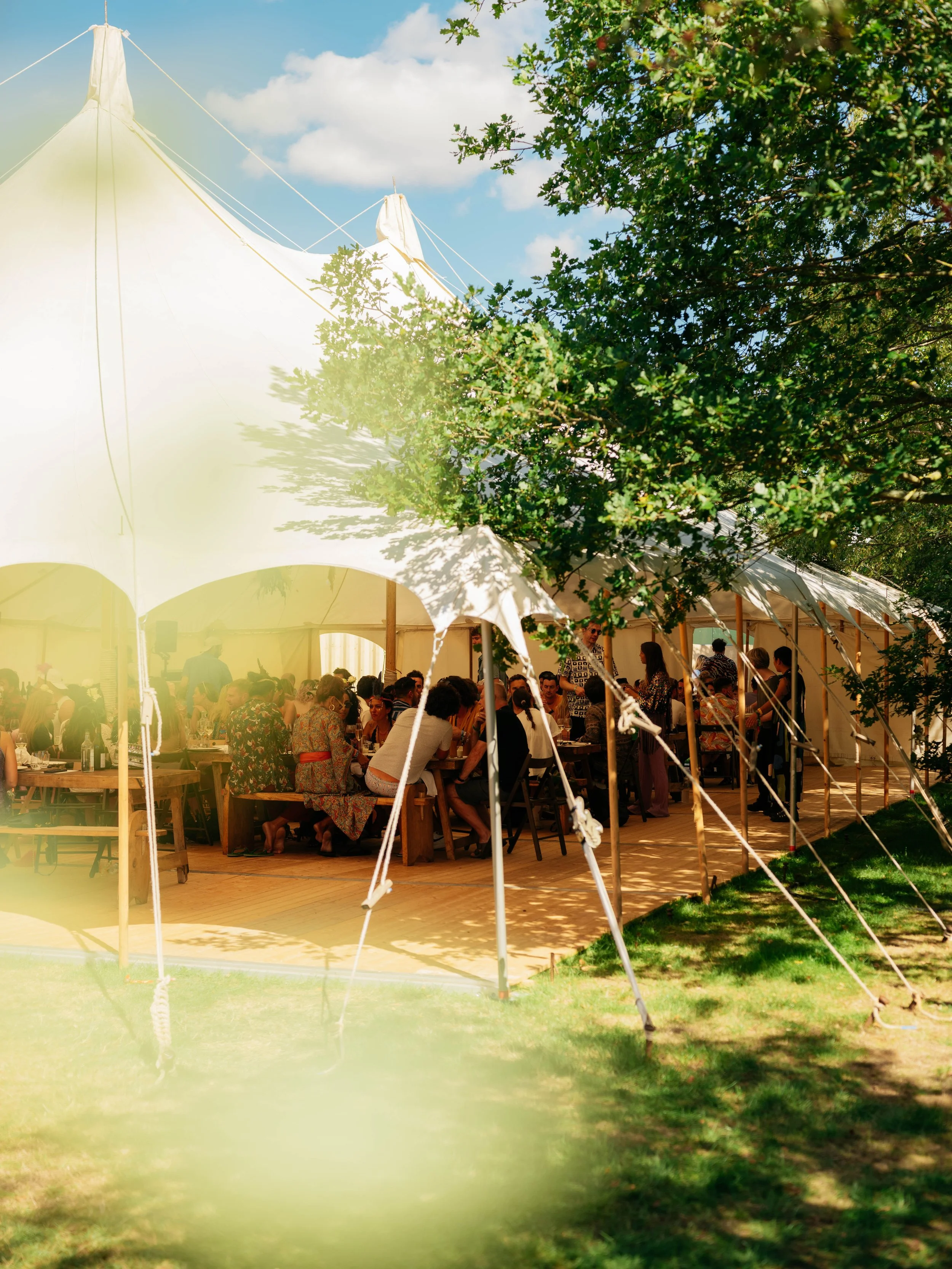 People gathered under a large white event tent, dining at a festival, with green trees and grass surrounding the area on a sunny day.