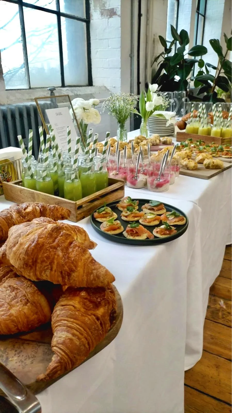 A table set with assorted pastries, finger foods, and beverages, including croissants, canapés with salmon and herbs, glass bottles of green juice with striped straws, small jars with pink desserts, and floral arrangements in vases, in a bright, industrial-style room with large windows.