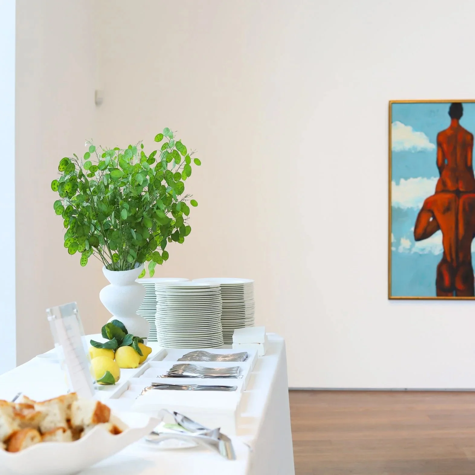 A buffet table with a green leafy plant in a white vase, stacked plates, lemons, silverware, and bread in a white dish. A colorful framed painting of a seated figure is on a beige wall in an art gallery.