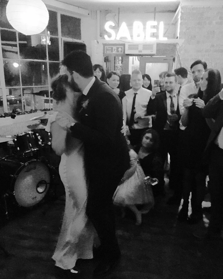 A black and white photo of a wedding reception dance floor with a bride and groom dancing in the foreground, surrounded by guests, and a drum set on the side.