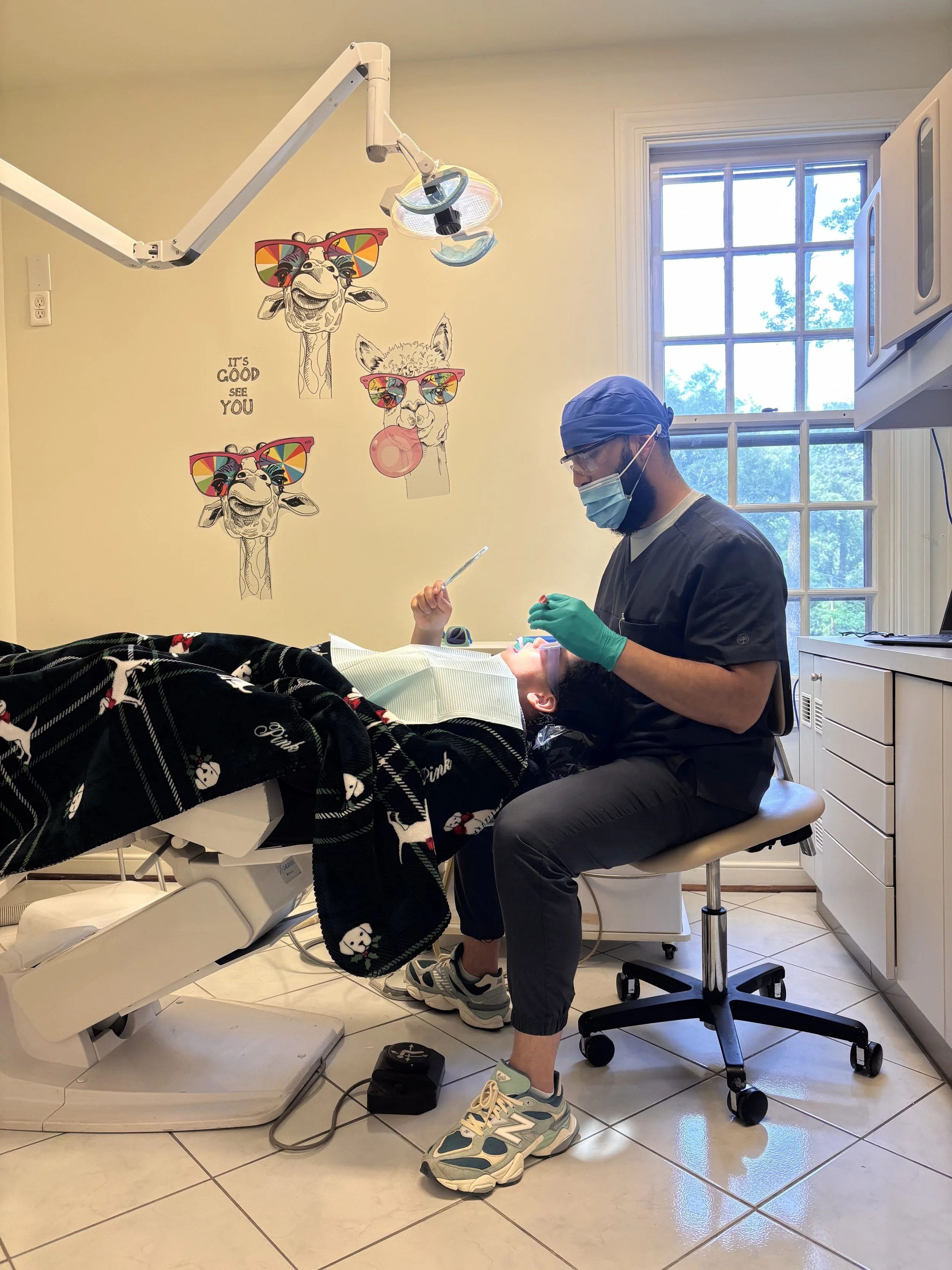 A dentist wearing a blue surgical cap, glasses, face mask, and gloves is sitting on a stool, working on a patient in a dental office. The patient is lying in a dental chair covered with a black blanket featuring white dog illustrations. The room has a window, dental equipment, and wall art of giraffes and a dog wearing colorful sunglasses, with the phrase "It's good to see you".