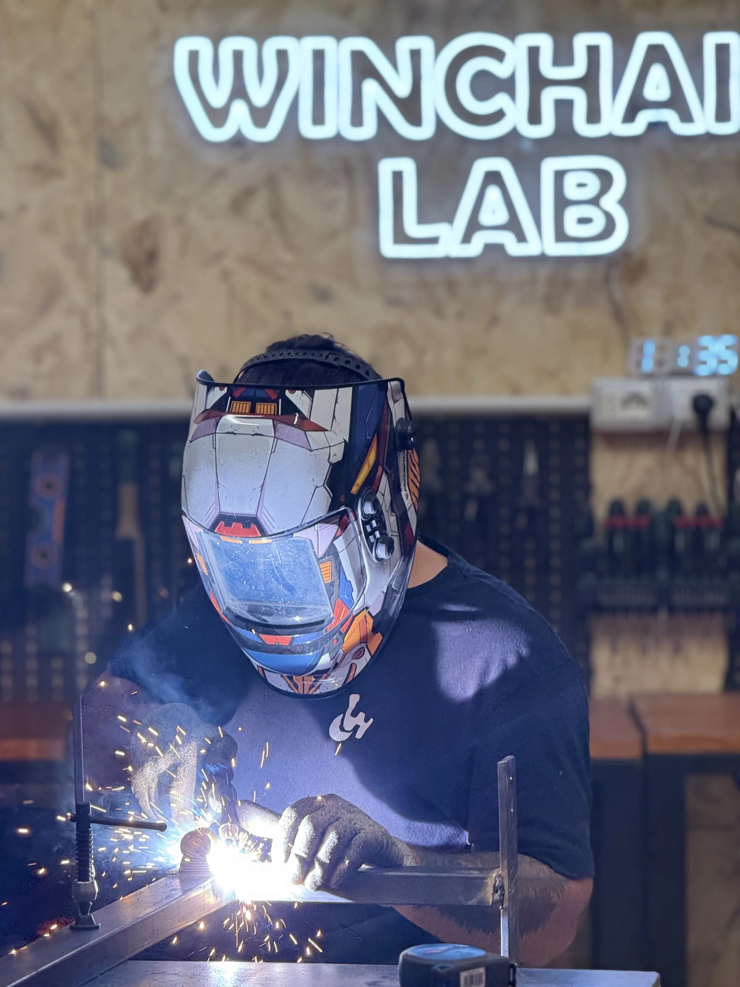 Un fabbro che indossa un casco da saldatura mentre lavora sul metallo, con scintille che volano durante il processo di saldatura, in un laboratorio chiamato 'WINGCHAN LAB'.