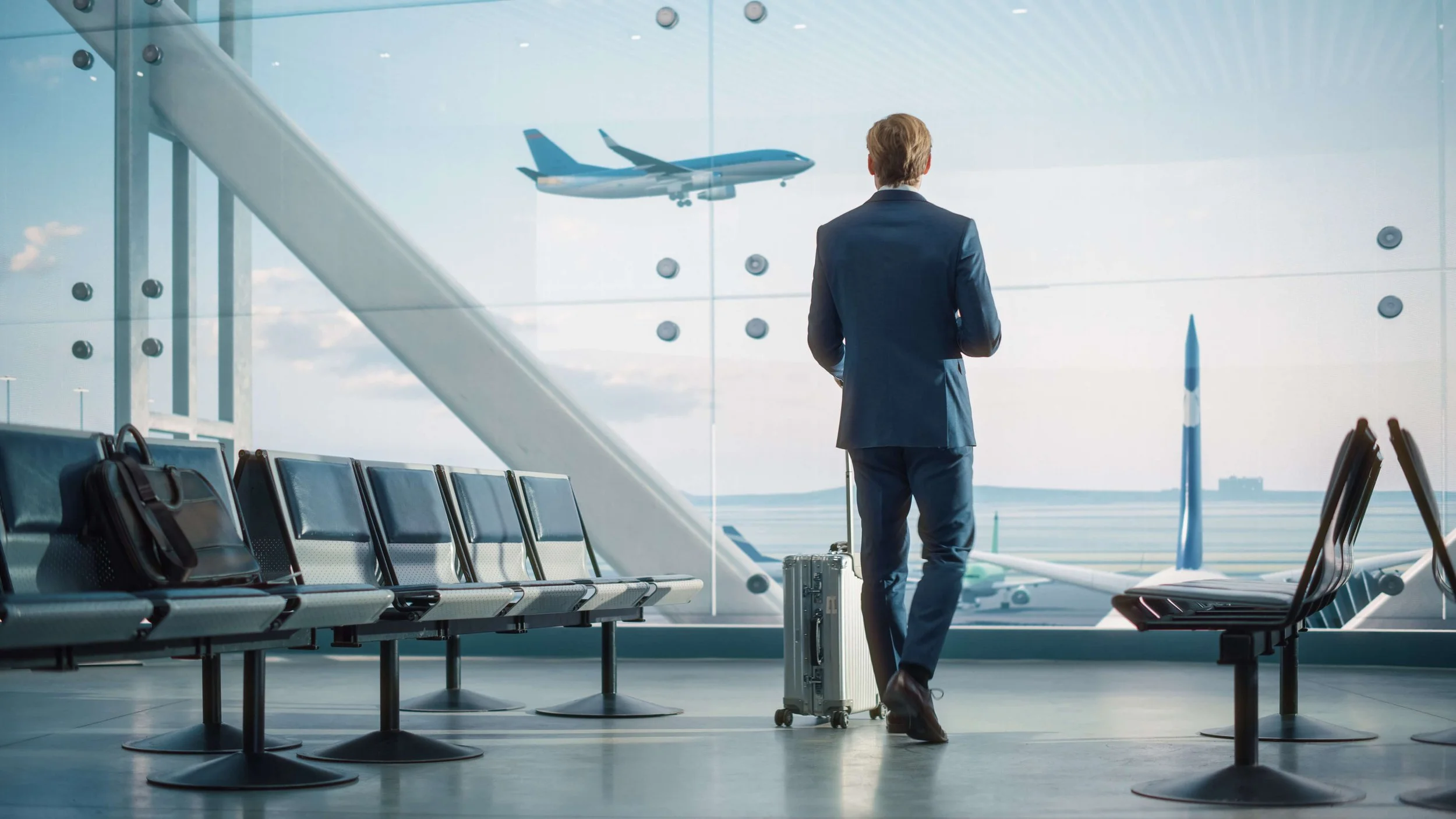 A man in a suit with a rolling suitcase standing in an airport waiting area, looking out the large glass windows at an airplane flying outside.
