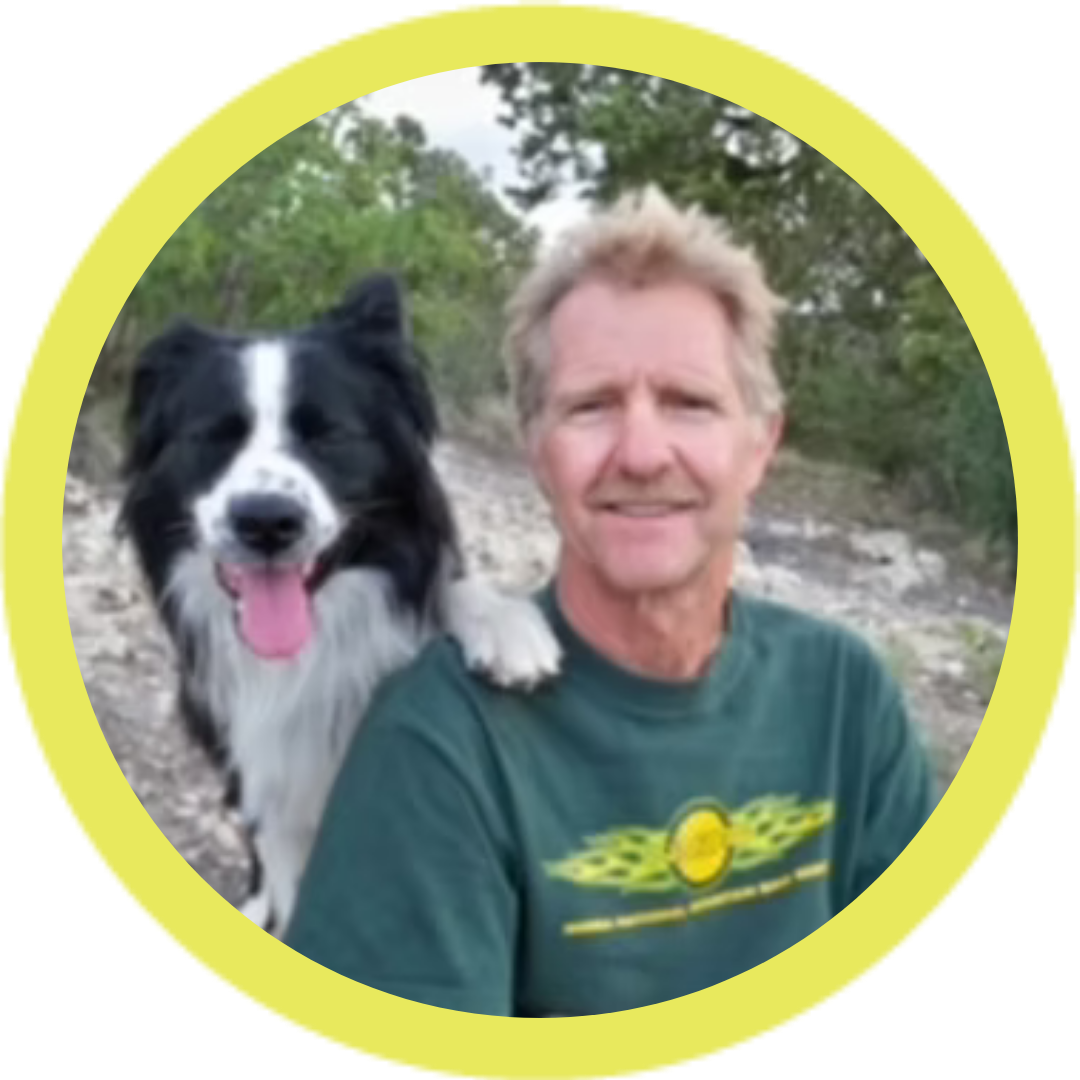 A man sitting outdoors with a black and white dog beside him, both smiling. The background shows trees and a rocky ground. The image is encircled by a yellow border.