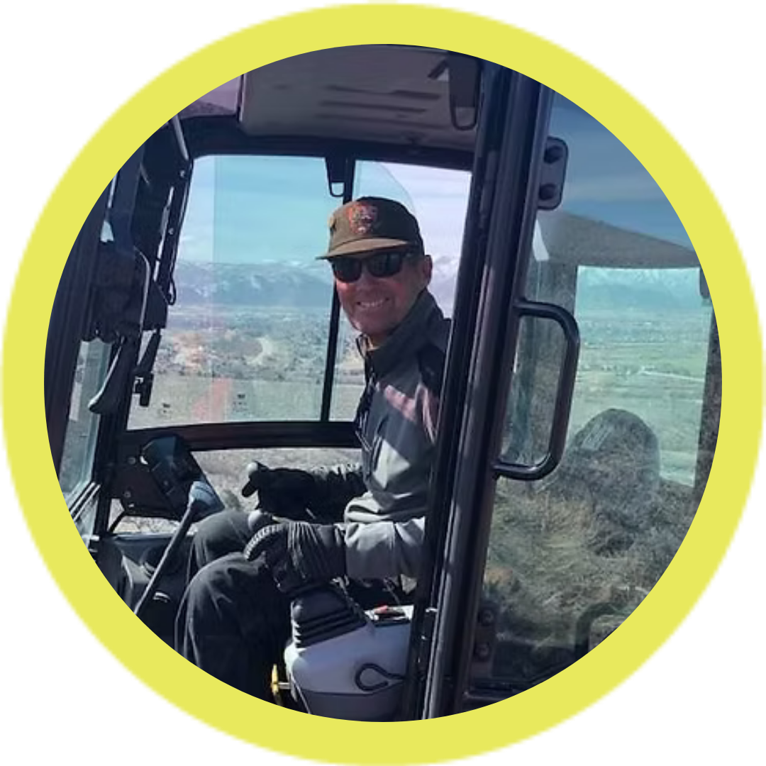 Person wearing sunglasses and a cap, sitting inside a construction vehicle with a view of mountains in the background.