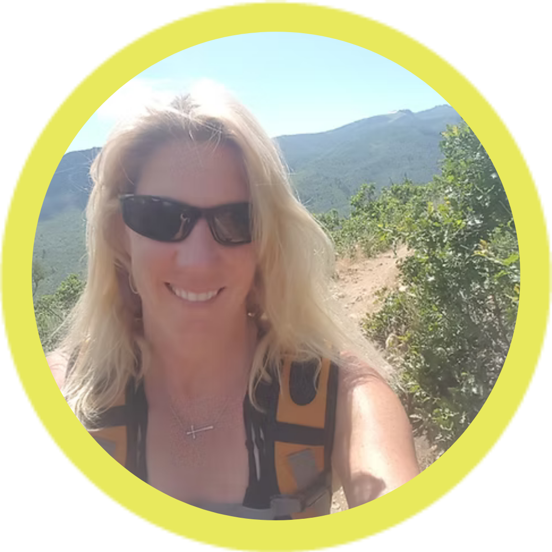 Person hiking on a mountain trail, wearing sunglasses and a backpack, with greenery and mountains in the background.