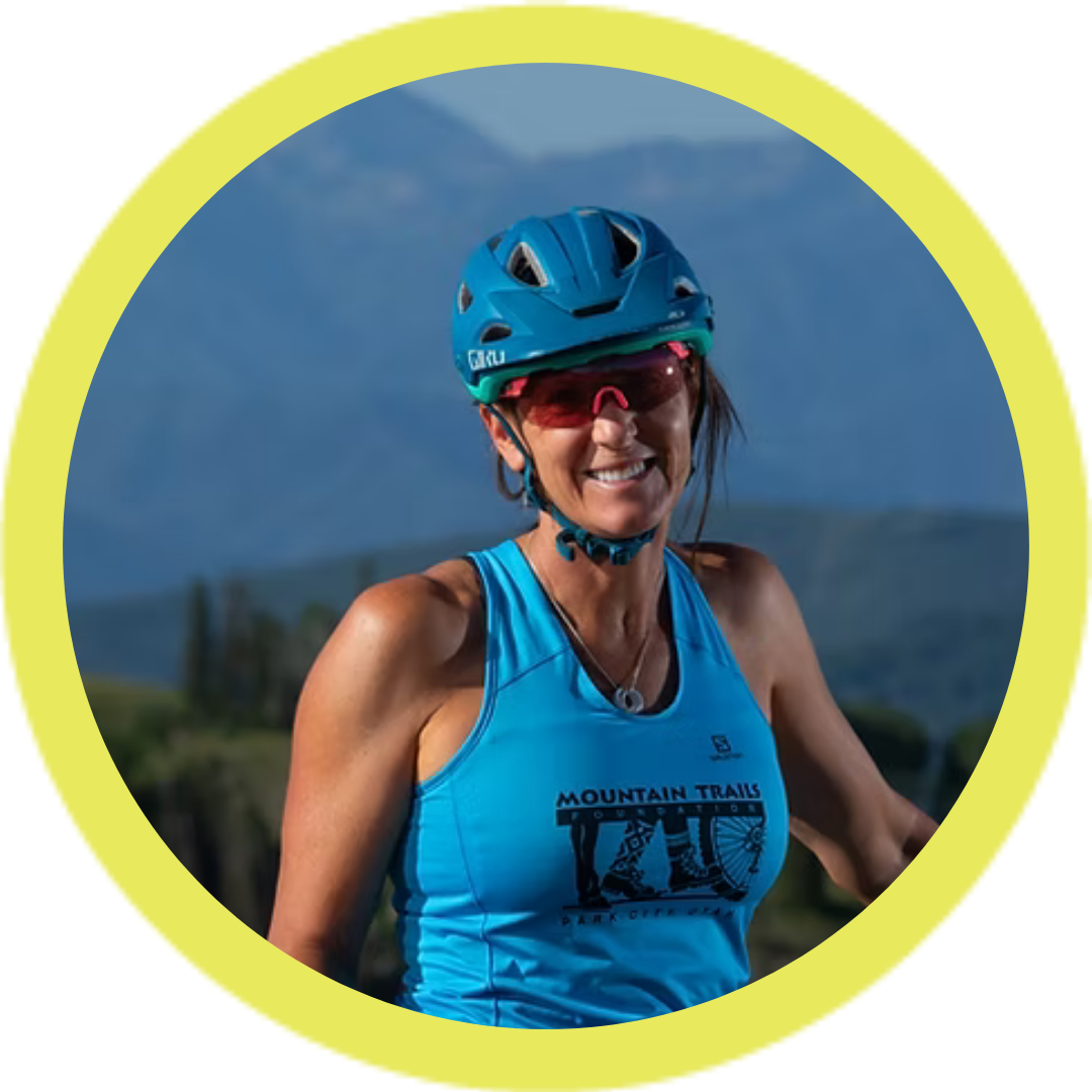 A woman wearing a blue helmet and sunglasses, smiling, with a sleeveless blue shirt that reads 'Mountain Trails Foundation' against a mountainous backdrop, framed by a yellow circle.