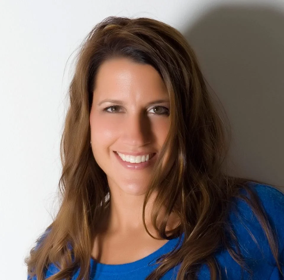 A woman with long brown hair smiling at the camera, wearing a blue top.