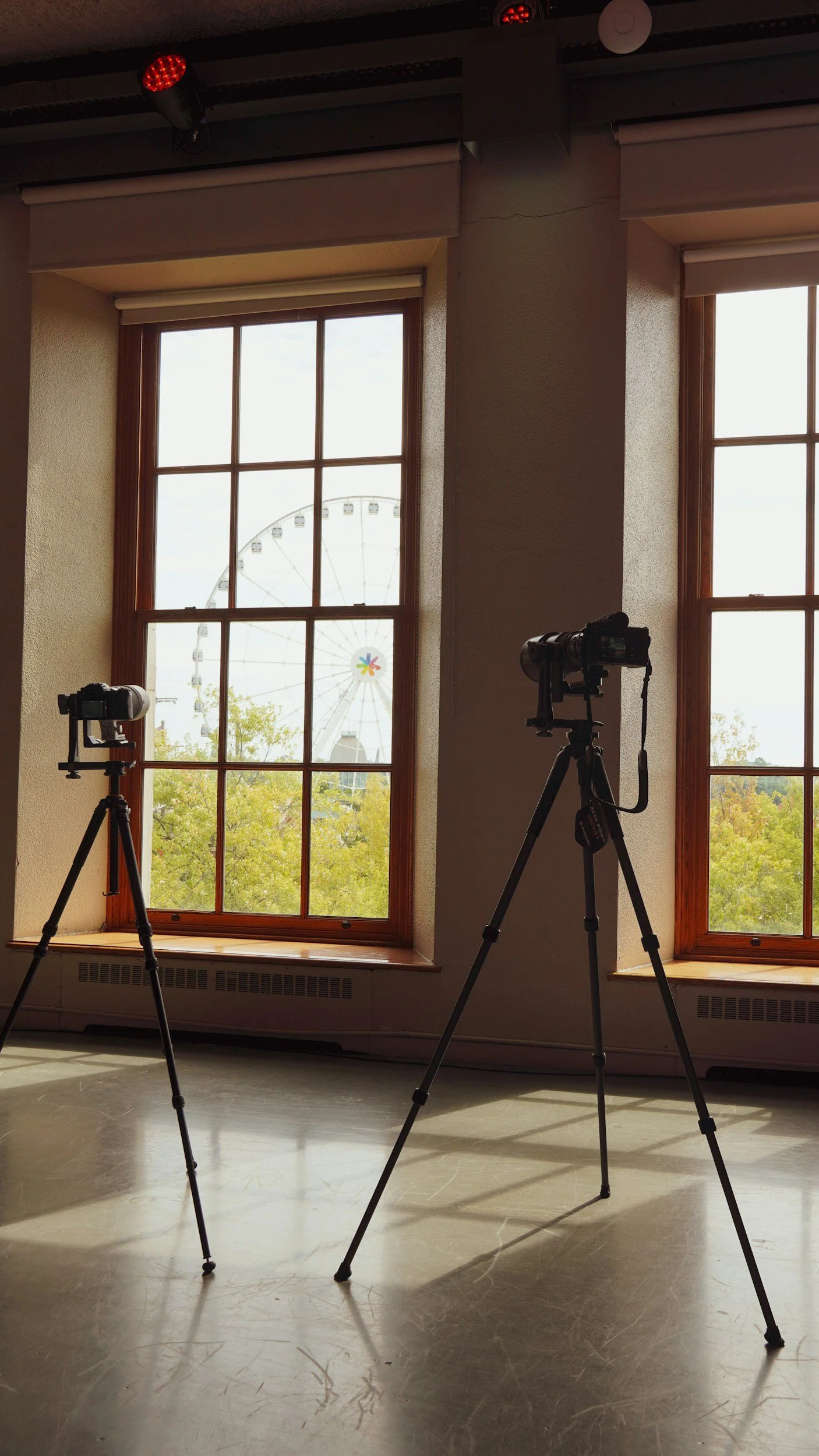 Two cameras on tripods set up inside a room in front of two large windows with a view of a Ferris wheel outside.