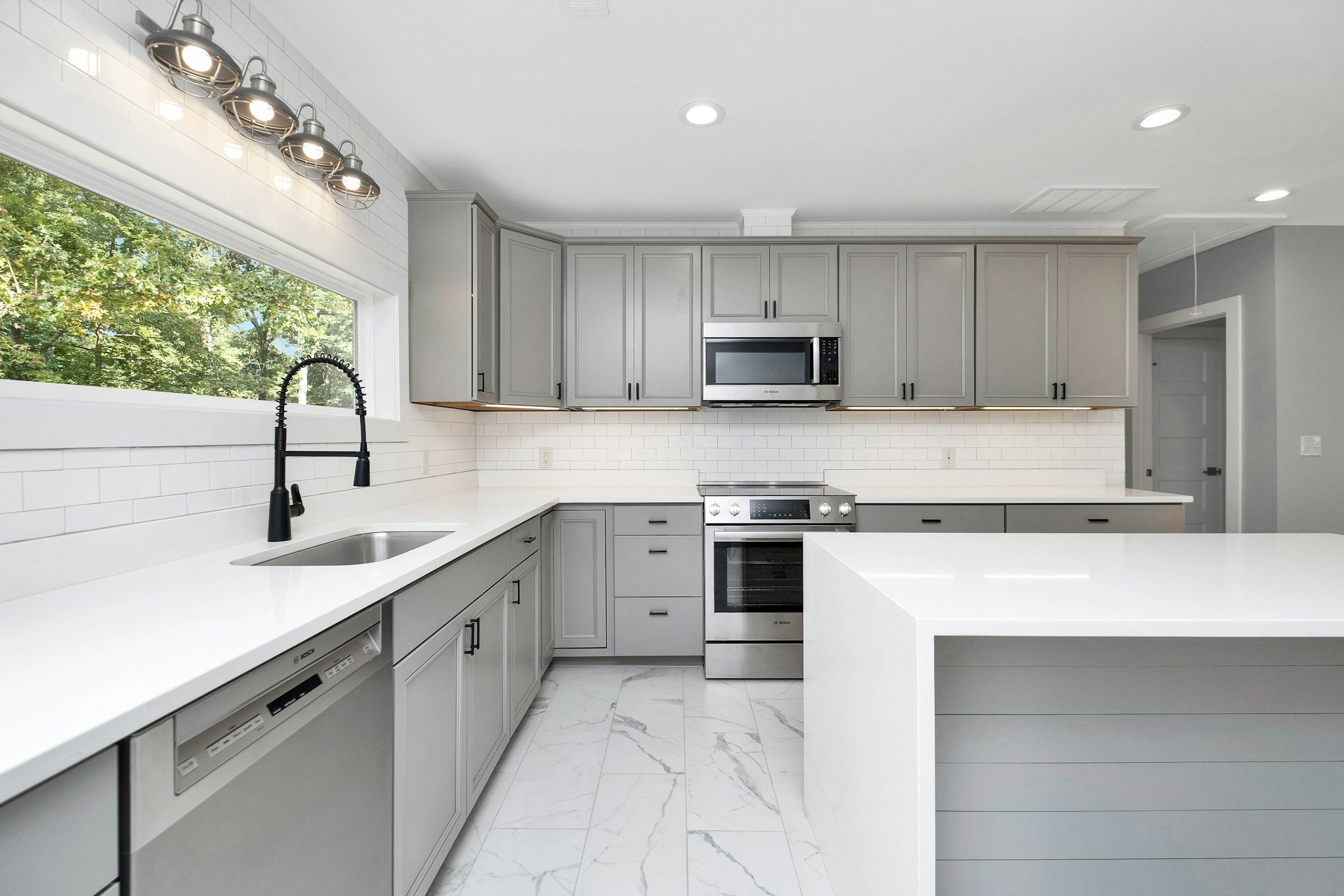 Modern kitchen with gray cabinets, white countertops, and stainless steel appliances, including a microwave and oven. There is a large window with a view of trees, and recessed lighting in the ceiling.