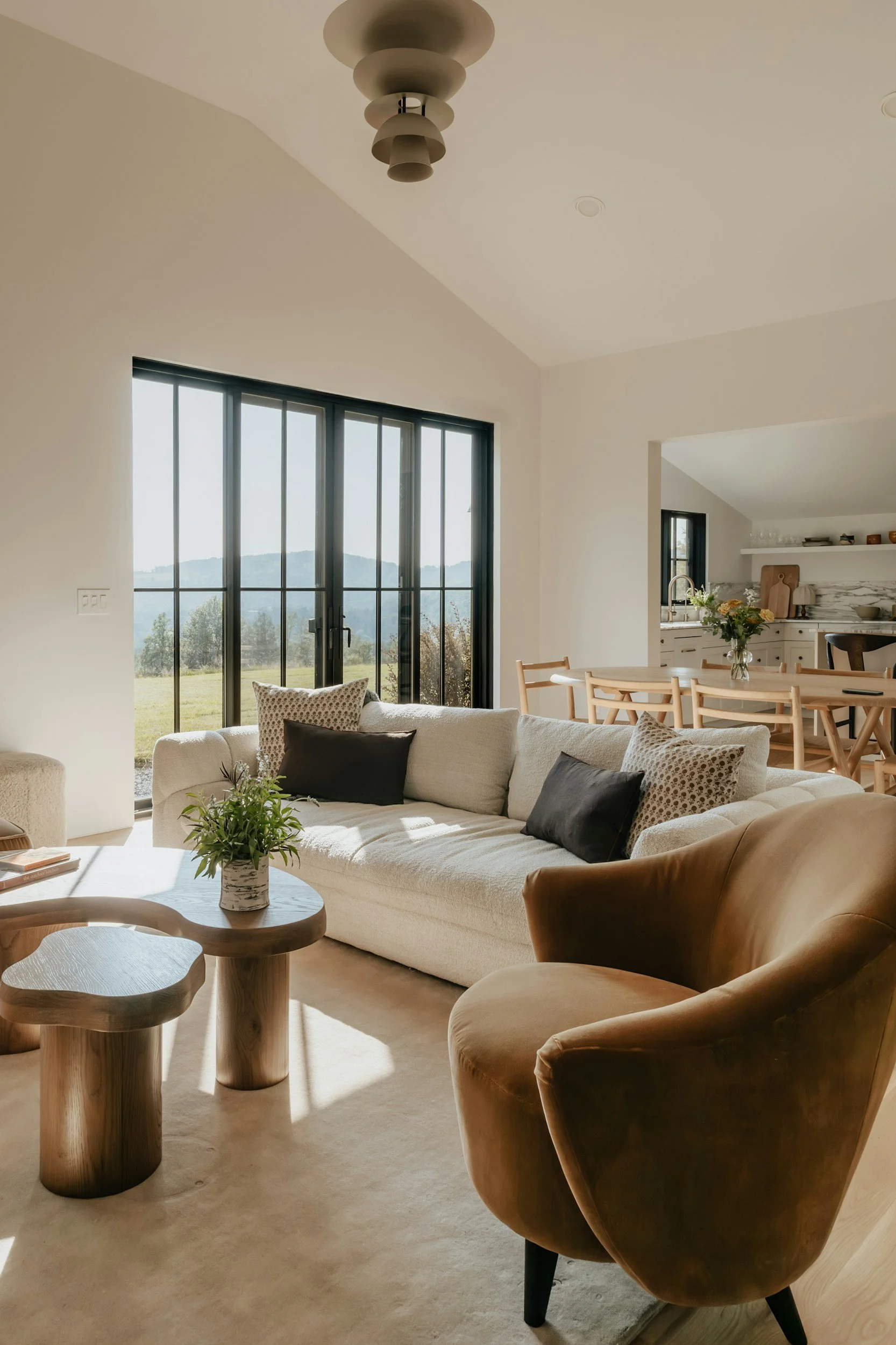 Living room with large glass sliding door, beige sofa with black and patterned pillows, wooden coffee table with a small potted plant, brown leather armchair, kitchen visible in the background with white cabinets and marble backsplash, dining table with chairs, and view of mountains outside through the glass door.