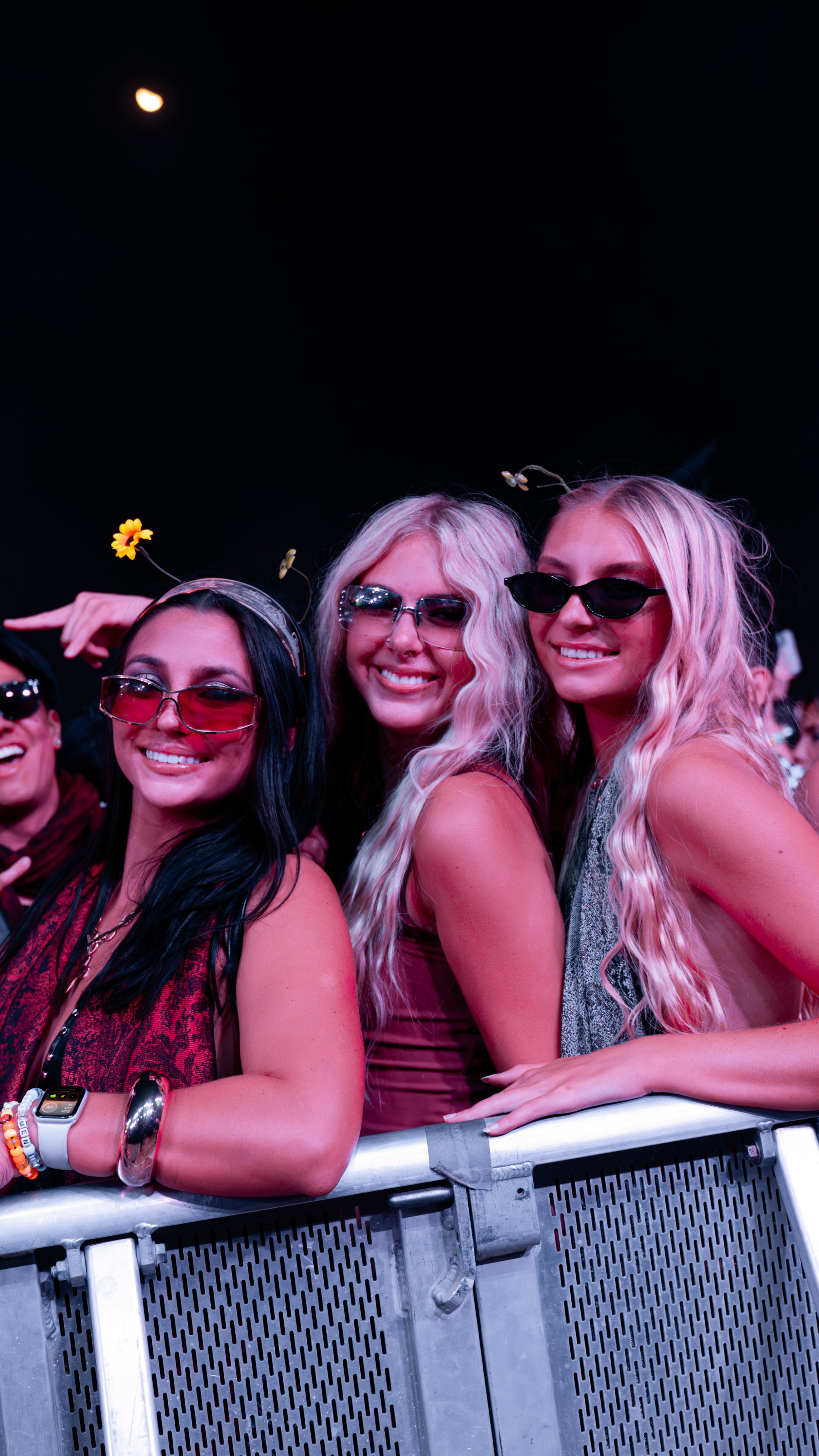 Three women at a concert or event, smiling behind a metal barrier, wearing sunglasses with yellow flowers in their hair.