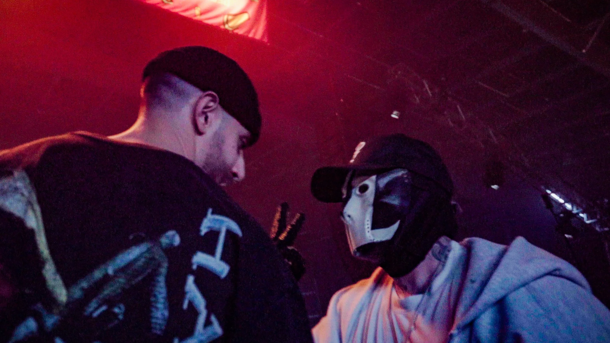 Two young men in conversation against a dark background with red and purple lighting. One is wearing a camouflage jacket and the other a gray hoodie and a hockey mask. 7L and Sisto.