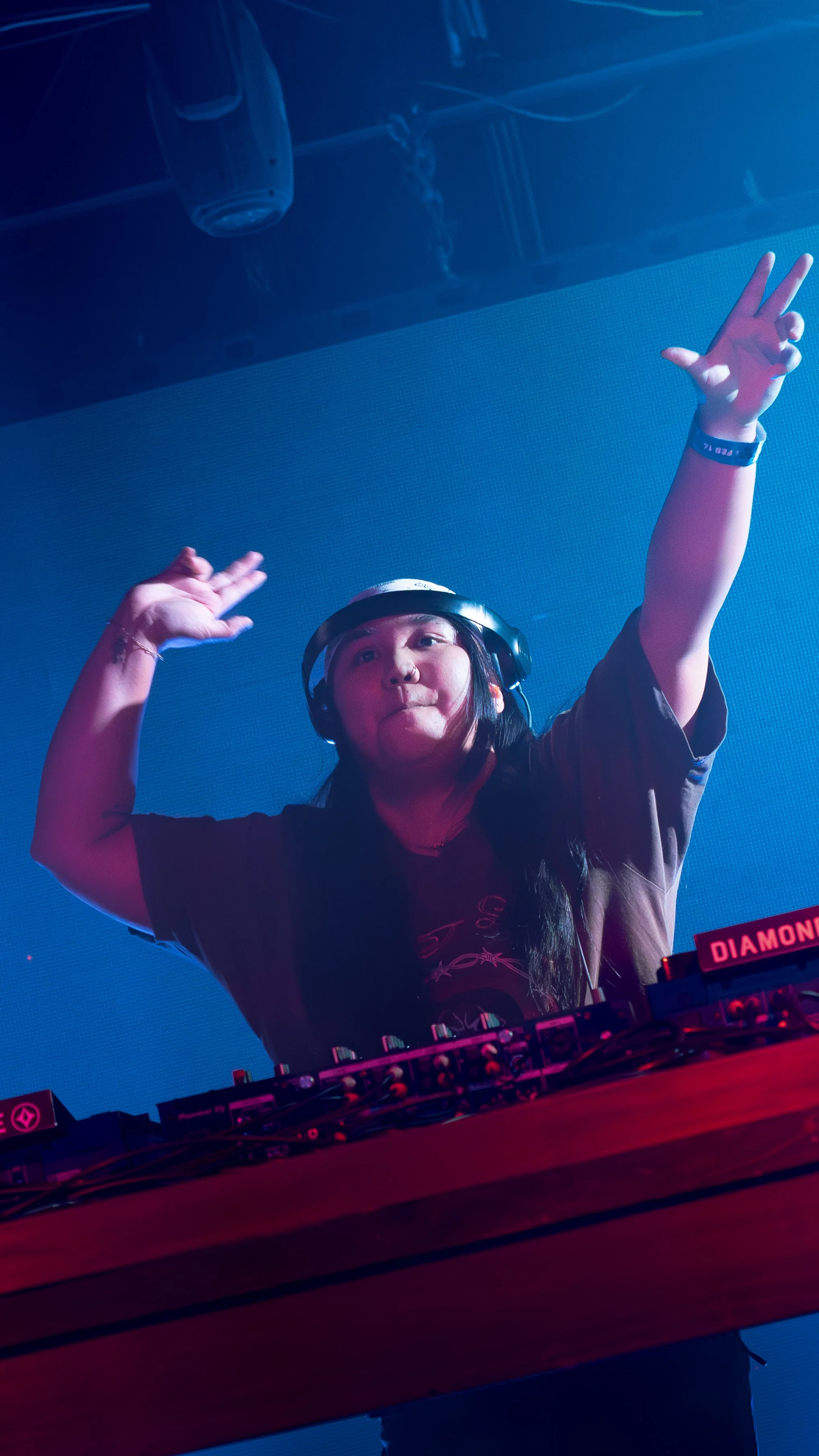 A female DJ with long dark hair wearing headphones, raising her hands in a dance pose, standing behind a DJ booth with highlighted controls, in a dark, vibrant environment with blue and pink lighting.