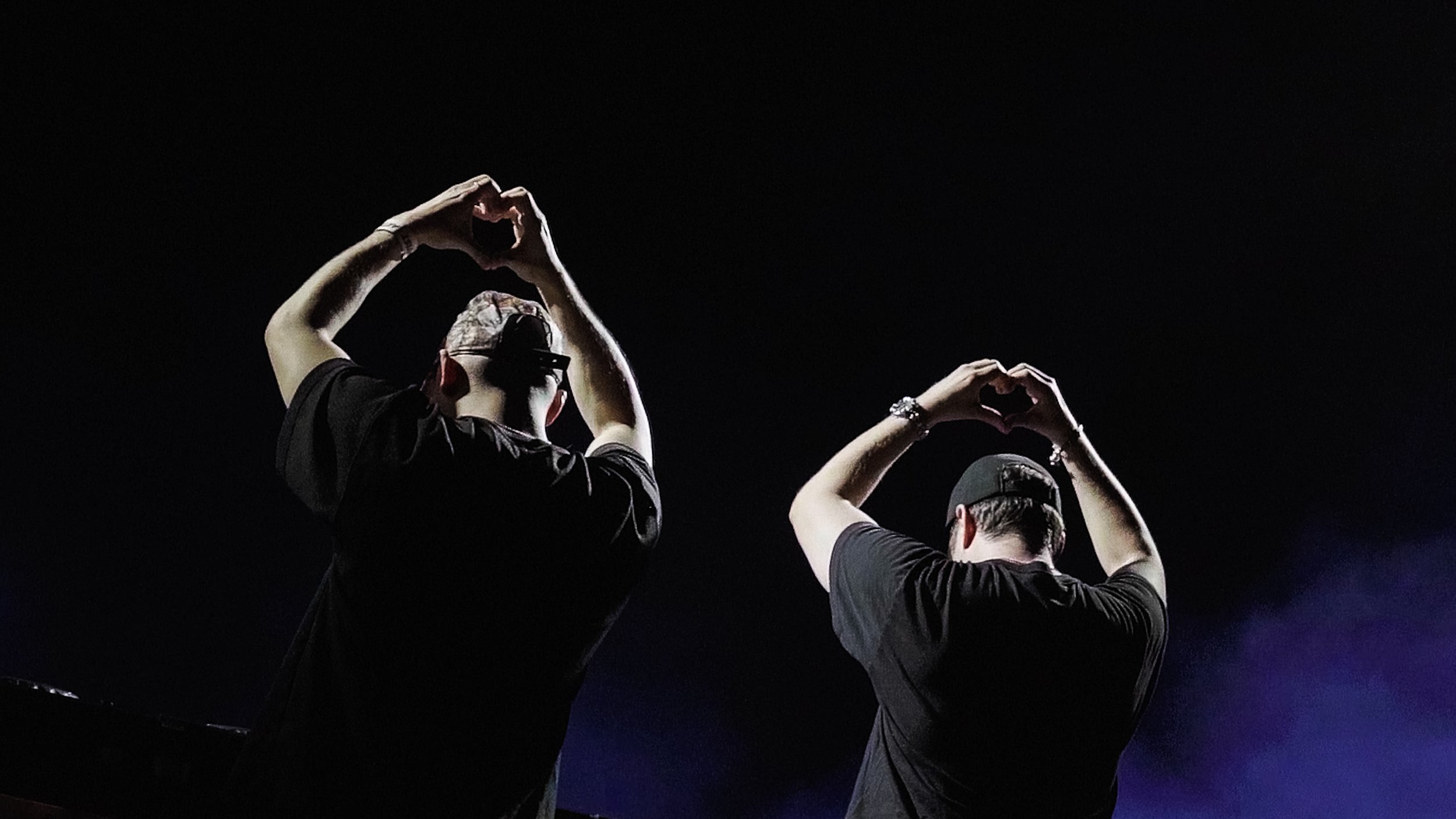 Two individuals in dark clothing and caps are making heart shapes with their hands against a dark background, illuminated from below. Slander.