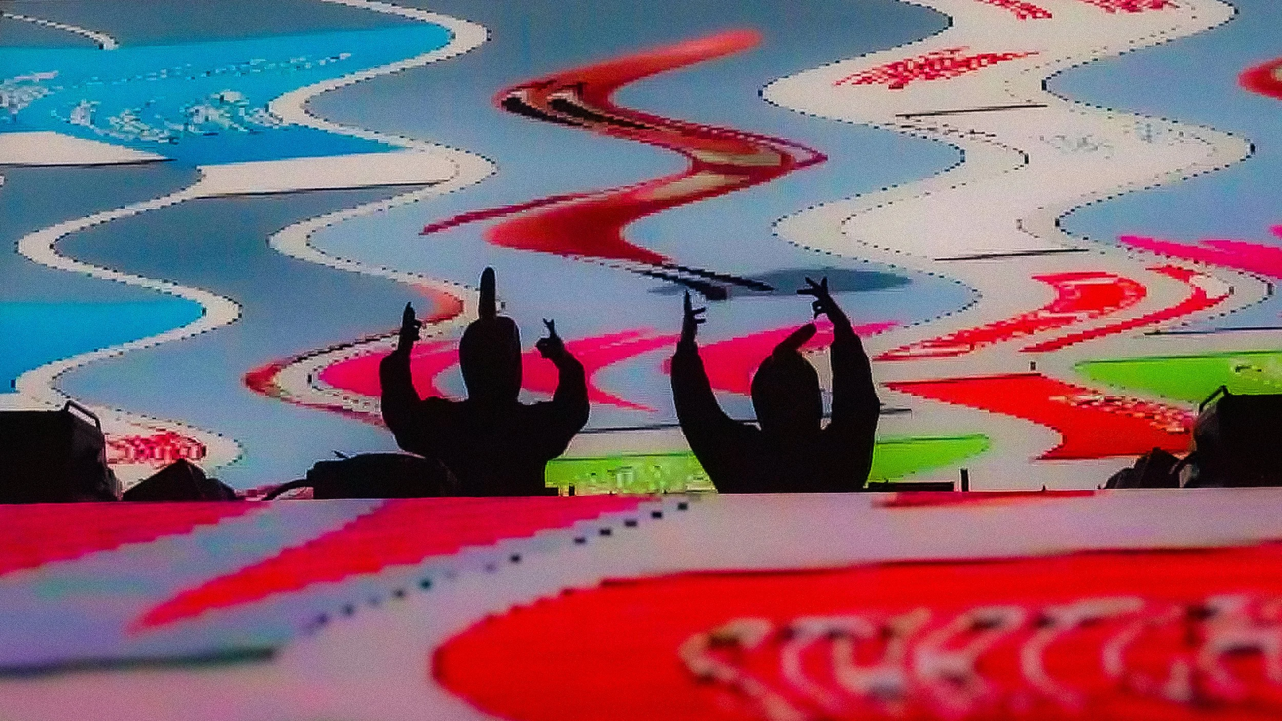 Silhouetted audience members at a concert or performance raising their hands with peace signs, with colorful abstract graphics on a screen behind them. Richard Finger.