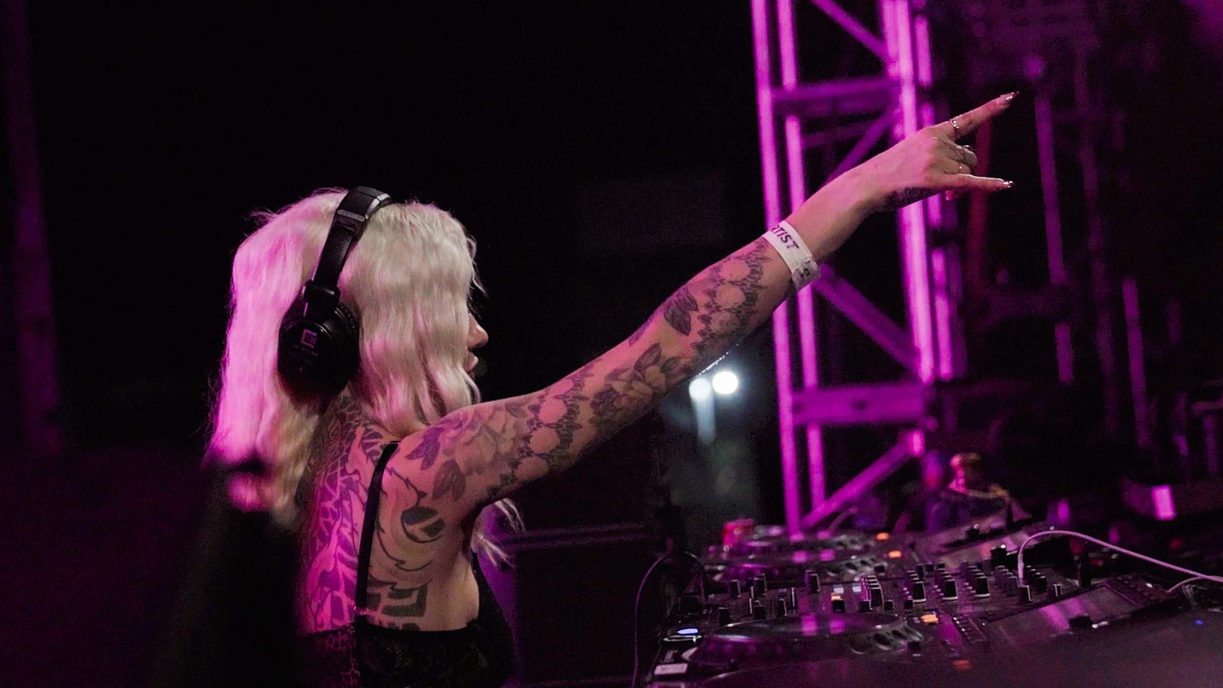 A female DJ with long blonde hair, wearing headphones, and covered in tattoos, is performing at a music event with purple stage lighting. Canabliss.