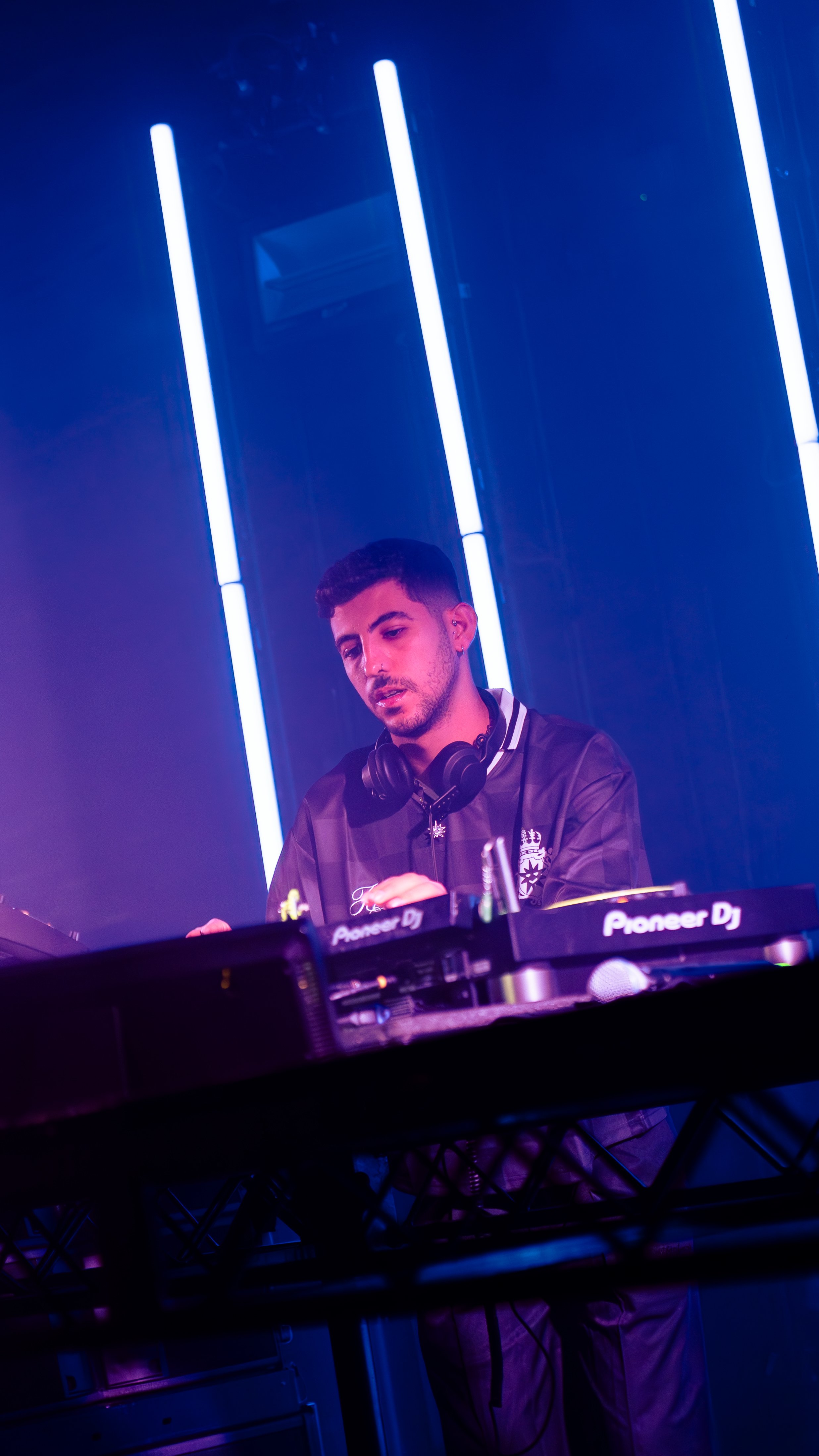 A DJ is performing at a nightclub, wearing headphones and focused on his equipment with bright vertical blue neon lights in the background.