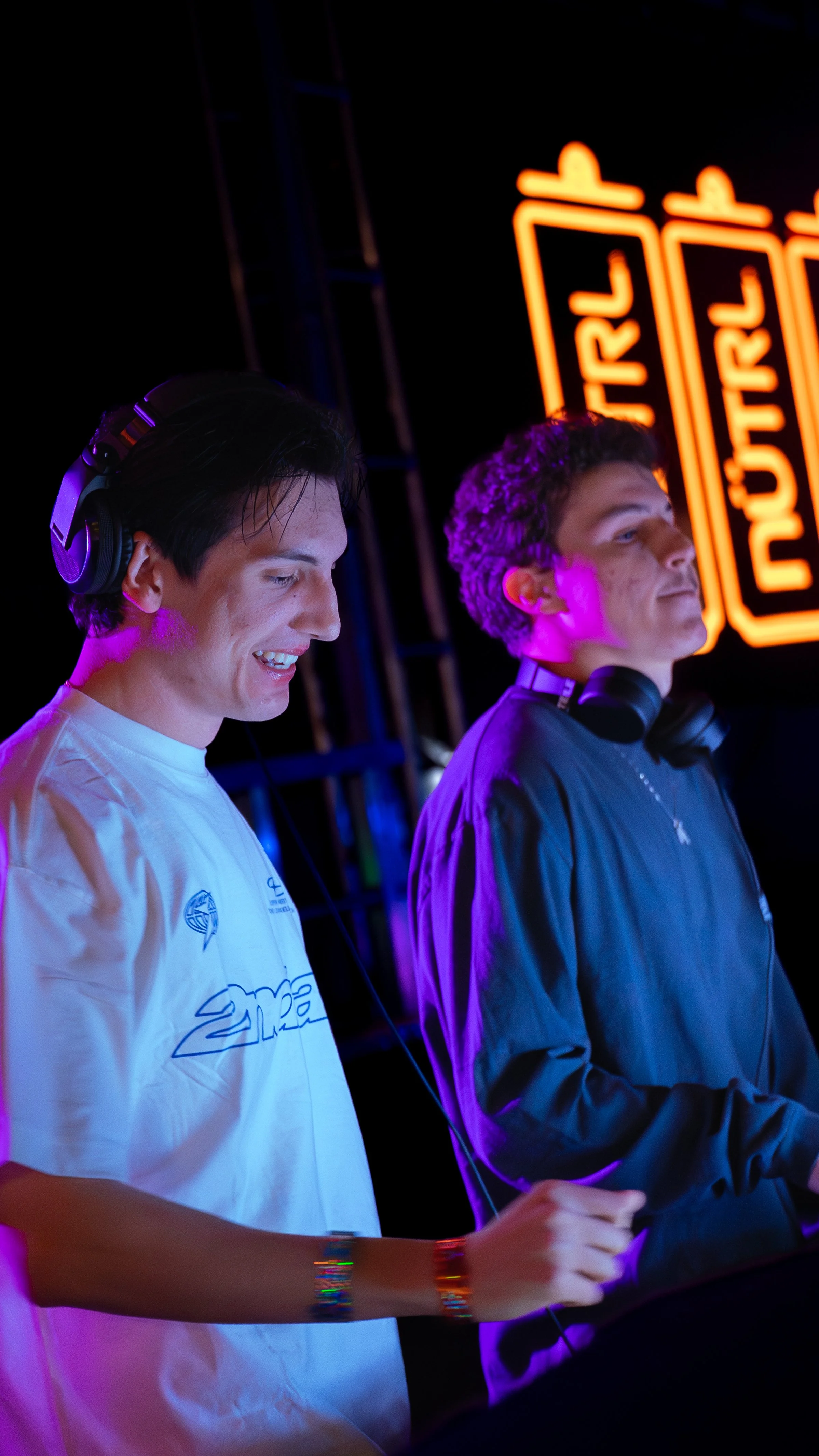Two young men wearing headphones, standing at a music event with a neon sign in the background. One is wearing a white T-shirt, smiling, and the other is wearing a black jacket, both looking towards the DJ.
