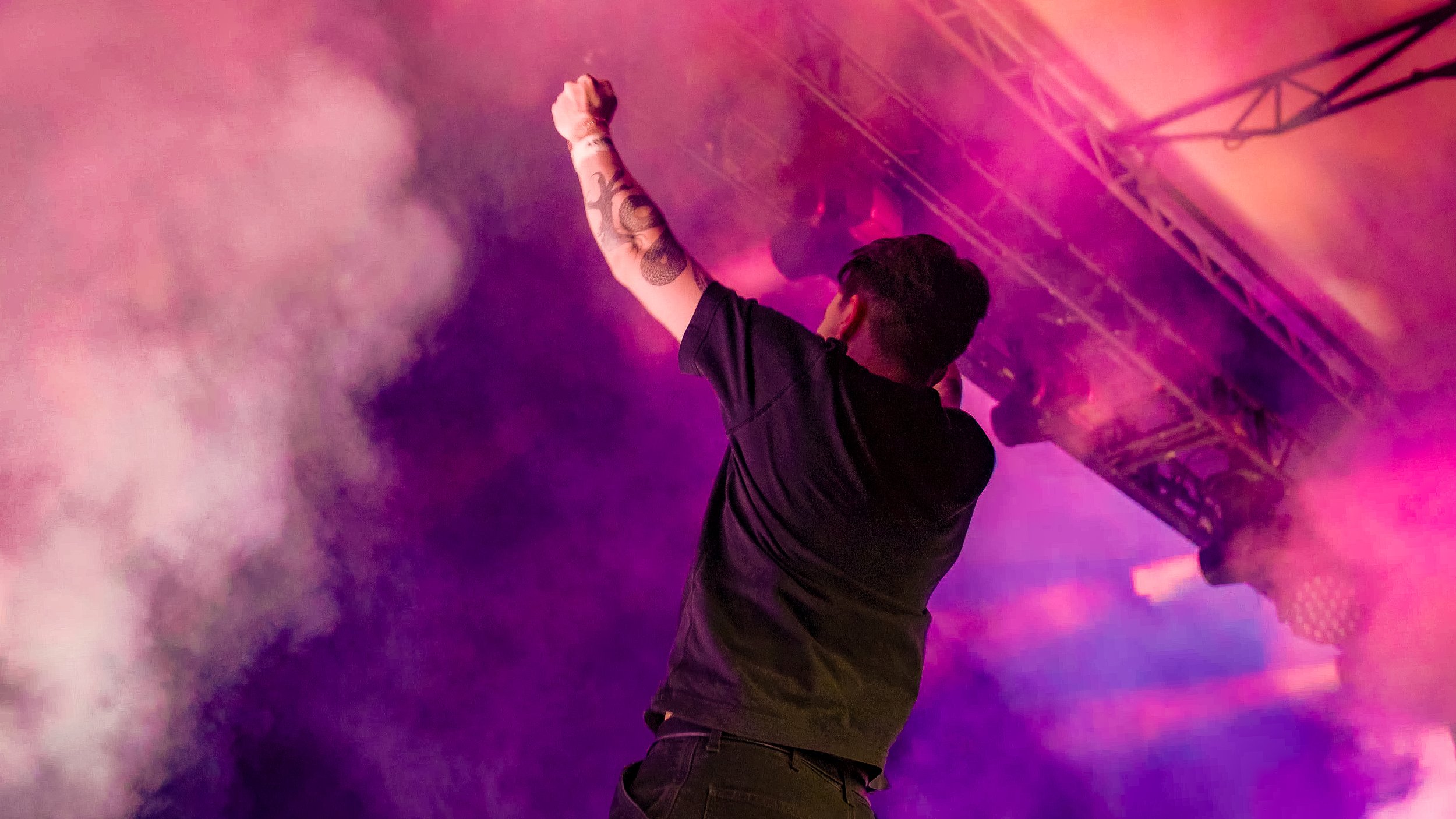 A person with dark hair and tattoos on their arm, raising their fist in the air at a concert or event filled with pink and purple lights and smoke. Eptic.