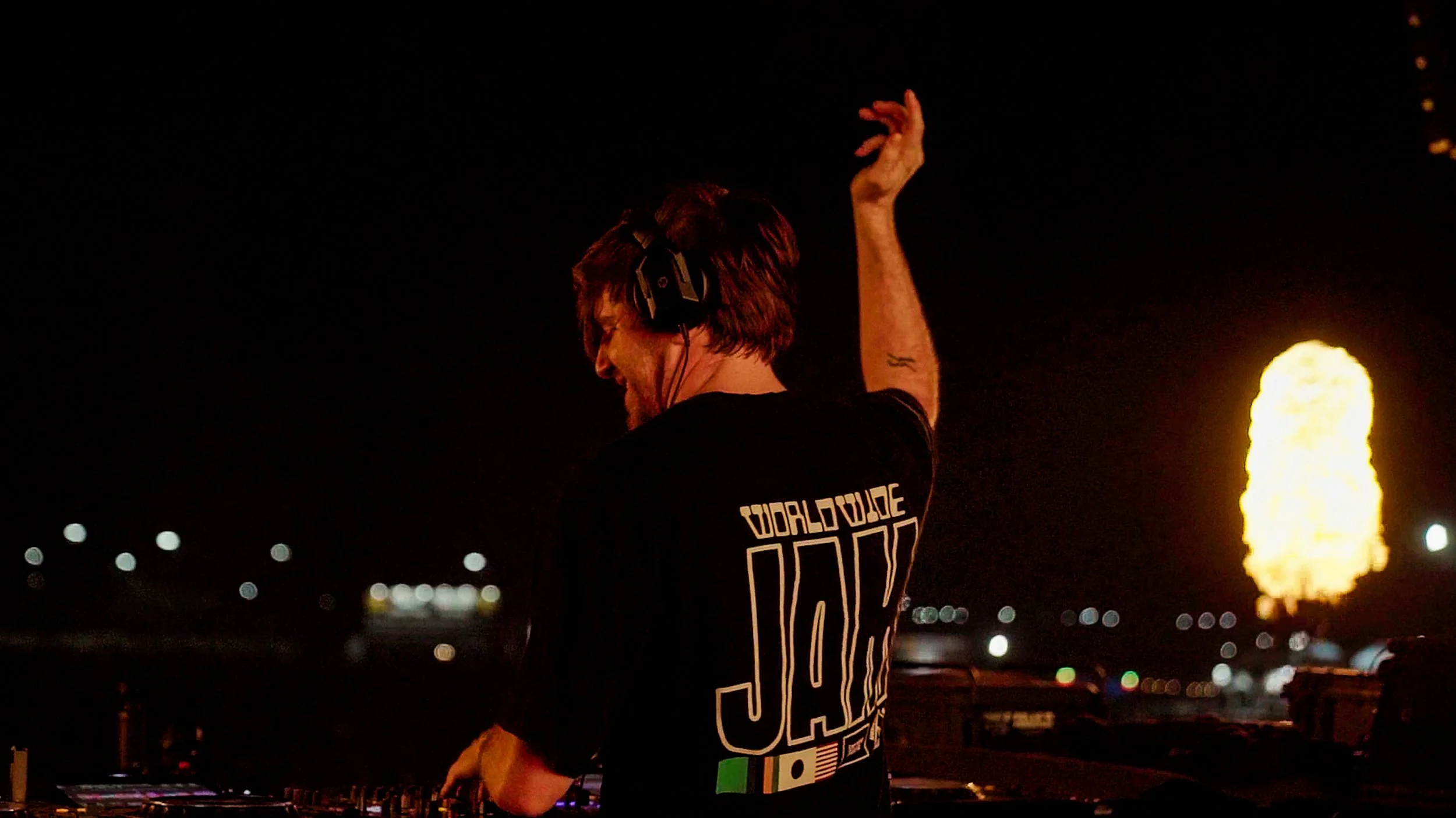 A person with red hair and headphones, wearing a black t-shirt, is DJing at night with fireworks in the background. Nghtmre.