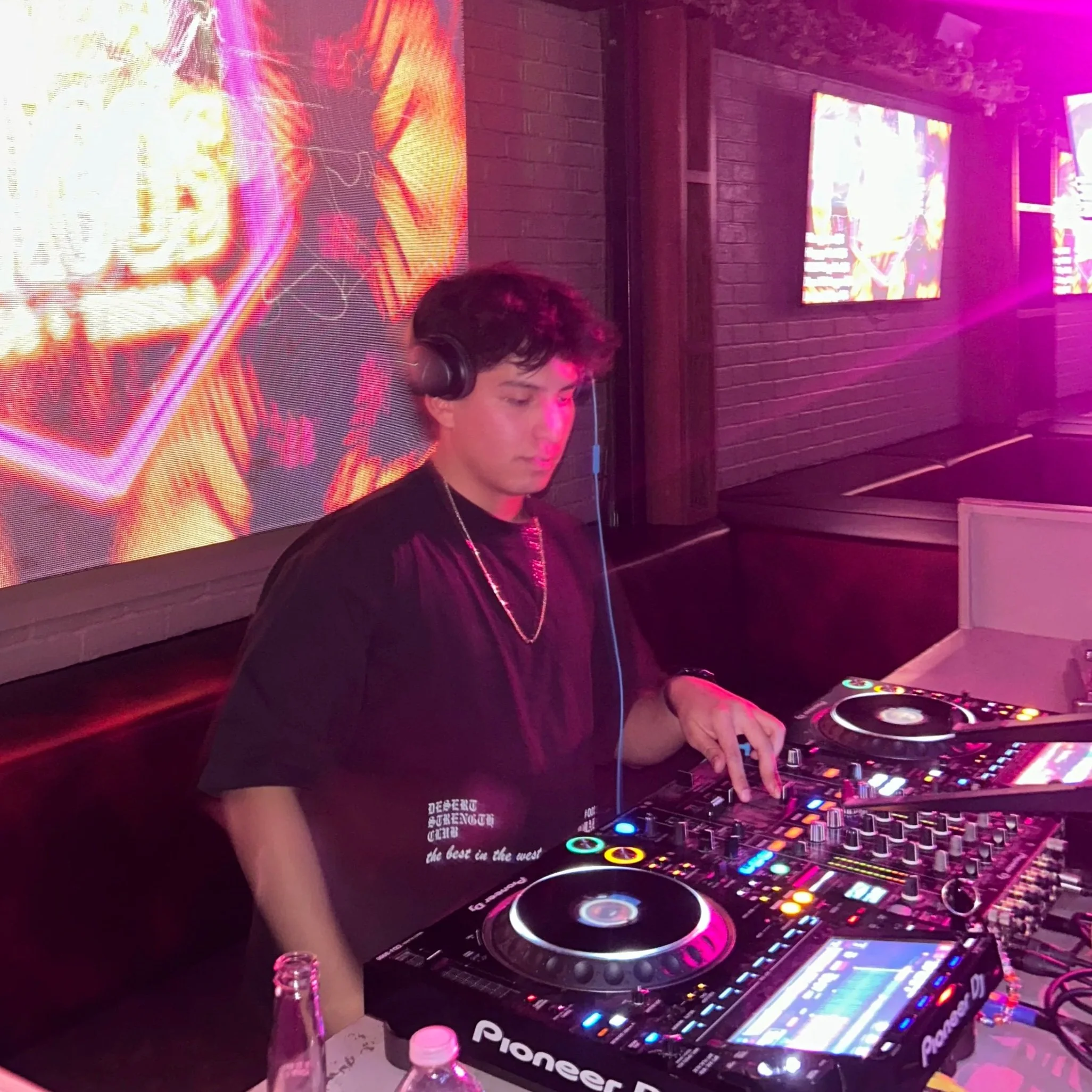 A young male DJ wearing headphones is operating a Pioneer DDJ controller at a club or party. The background features illuminated screens displaying vibrant, colorful visuals, with a brick wall and pink-purple lighting evident in the setting.