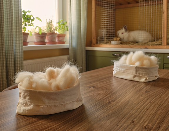 two fabric baskets hold harvested Angora wool white the rabbit rests in a clean cage nearby