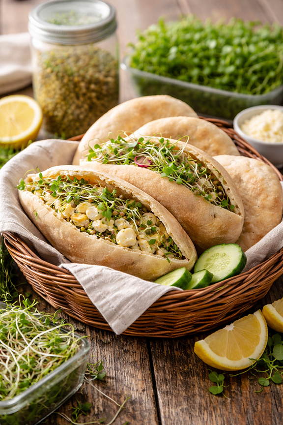Pita sandwiches in a basket with a linen cloth, a jar of sprouts, and microgreens