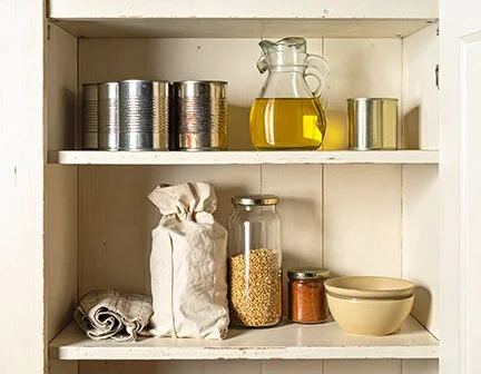 A small, open shelf pantry with cans, a cruet of oil, a bag of something, a jar of small pasta, a jar of sun-ripened tomatoes, and a bowl