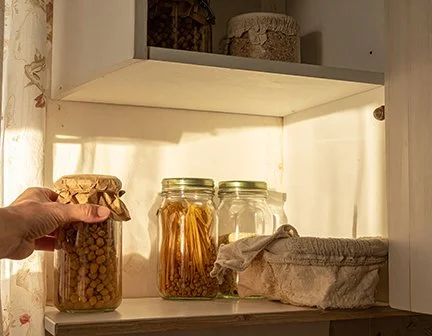 An open pantry shelf with jars of home canned goods and dry goods in jars. A hand is taking one jar off the shelf