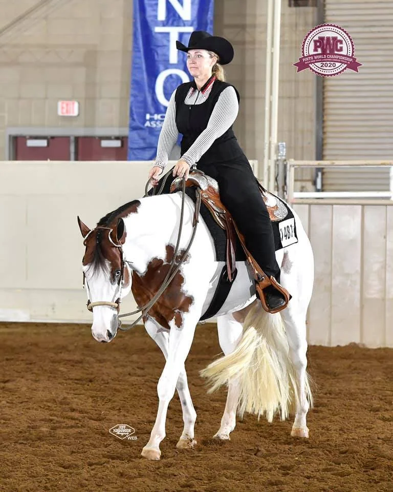 Woman riding a paint horse in an indoor arena during a horse show, wearing western attire with a backdrop featuring "PWC Paint World Championship 2020" logo.