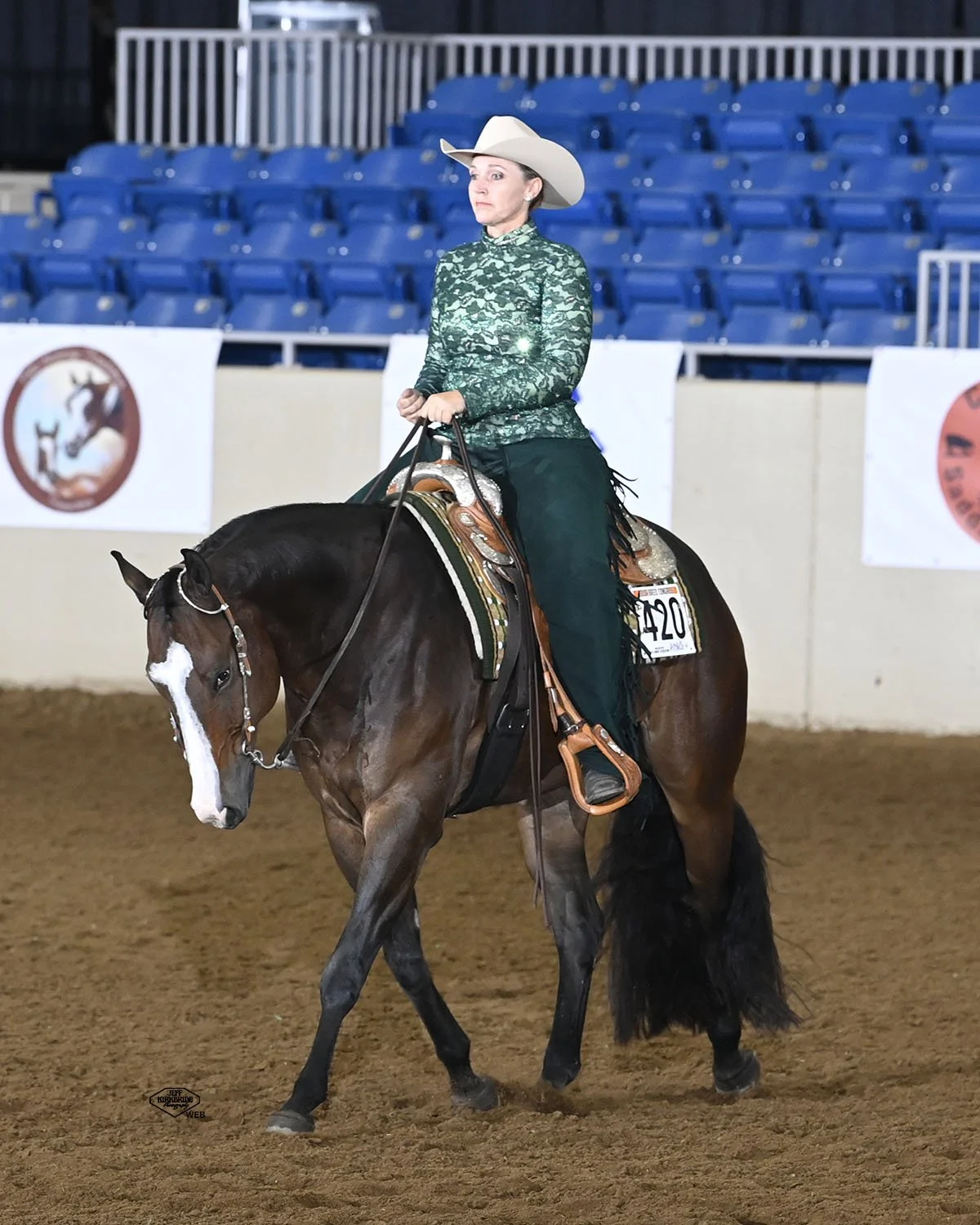 A woman dressed in Western attire, including a white cowboy hat and a green long-sleeve top, riding a brown horse in an indoor arena with blue seating and event banners.