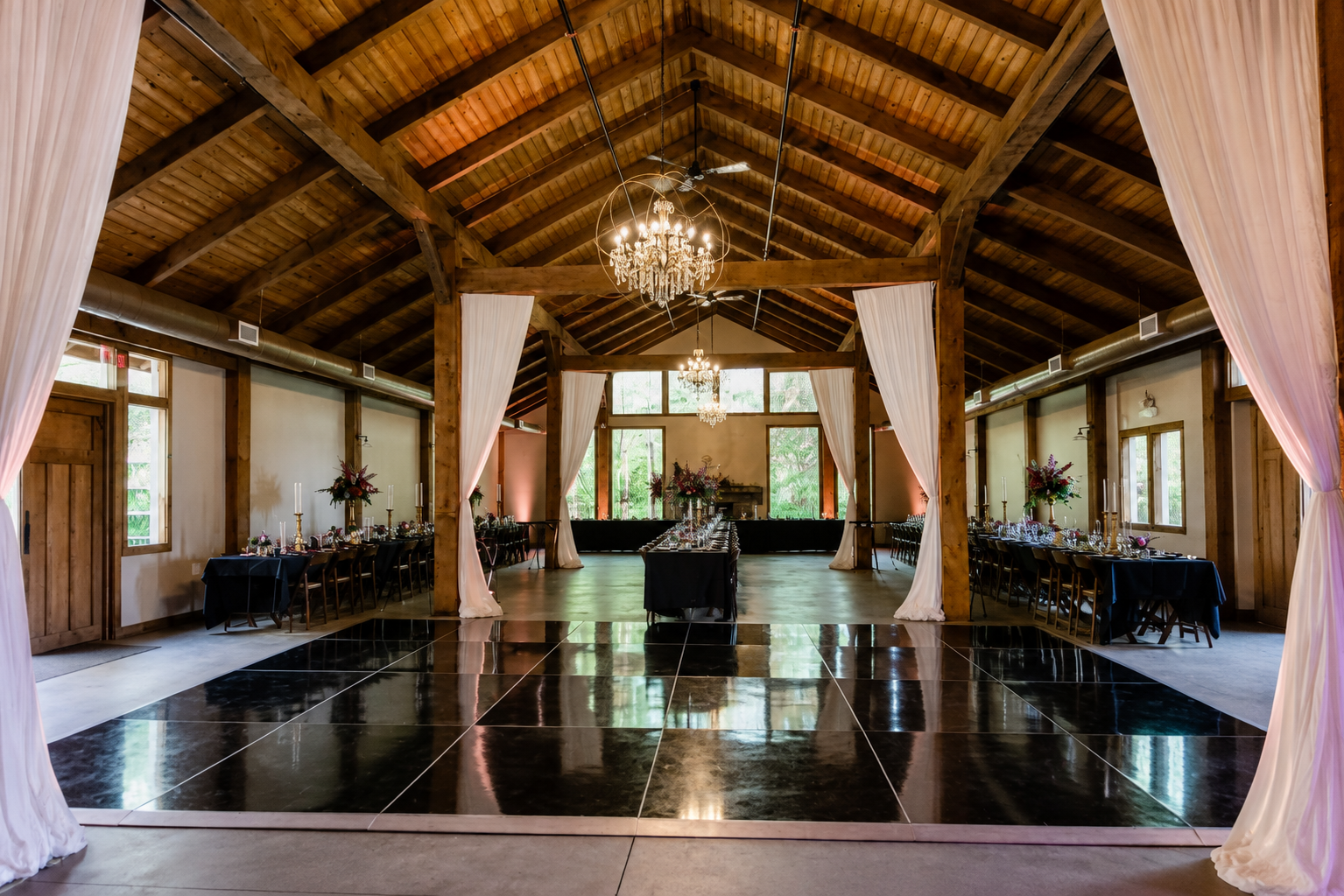 Elegant event hall with wooden beams, chandeliers, and tables set for a celebration, decorated with floral centerpieces and candles.