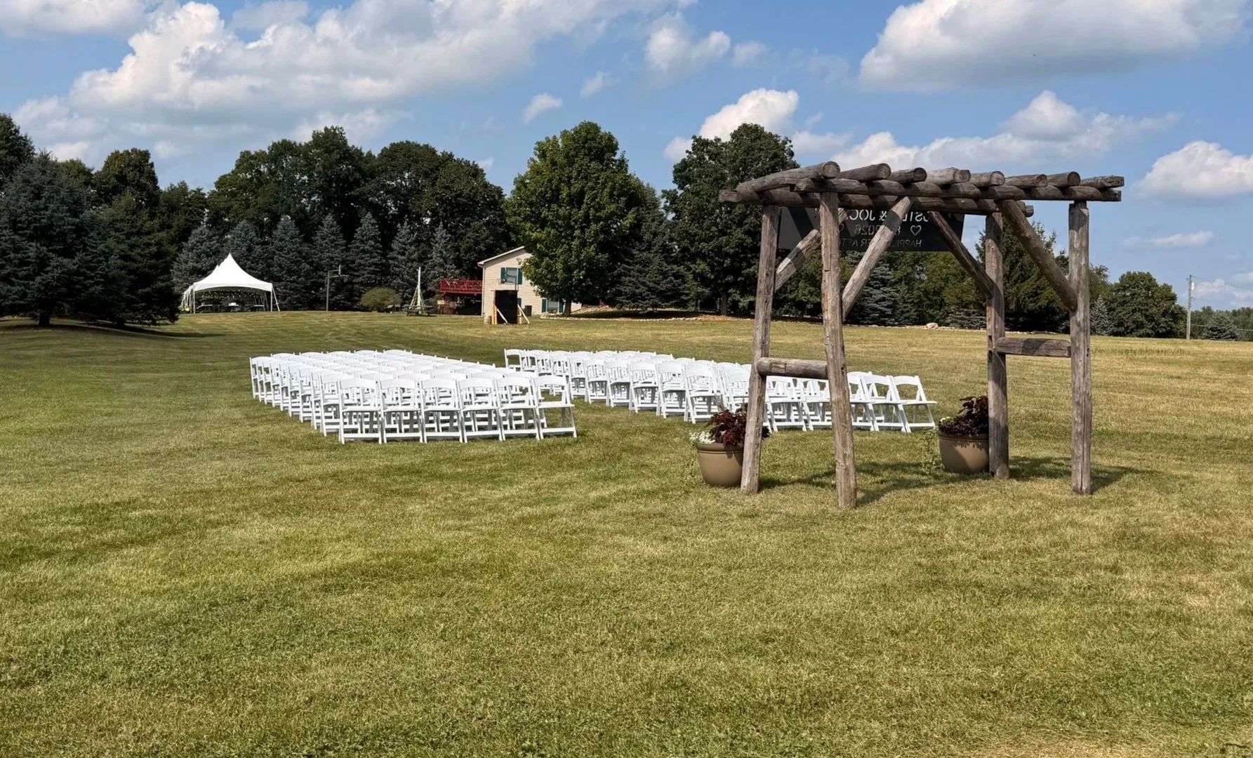 Outdoor wedding ceremony setup with white rental chairs, wooden arbor and event tents on a grassy venue.